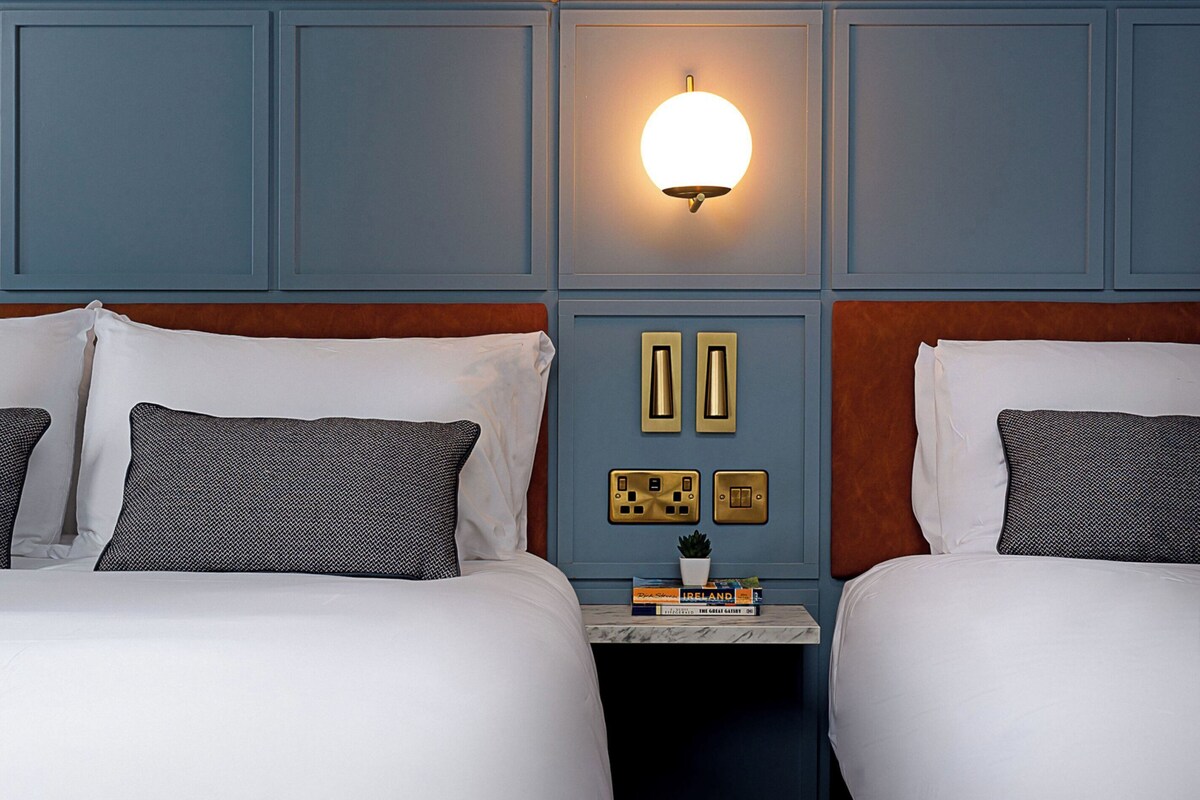 A hotel room with two beds, a nightstand with a lamp, and a wall-mounted light fixture.