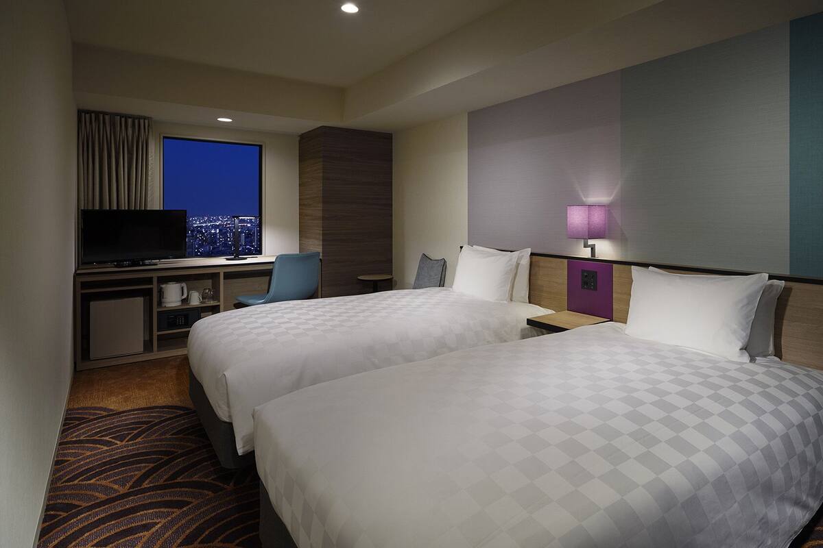 A hotel room with two beds, a desk with a TV, a city view, and a nightstand with a lamp.