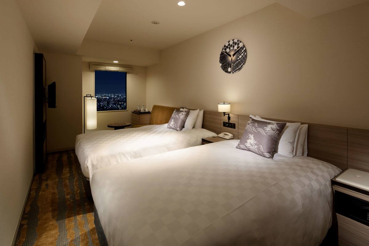 A hotel room with two beds, a nightstand with a lamp, a wall clock, and a window with a city view.