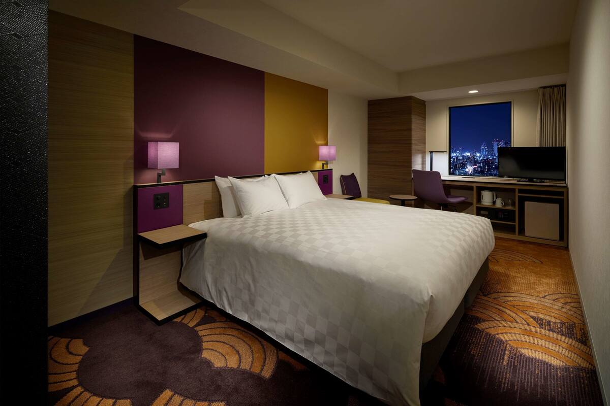 A hotel room with a large bed, a desk with a TV, and a city view.