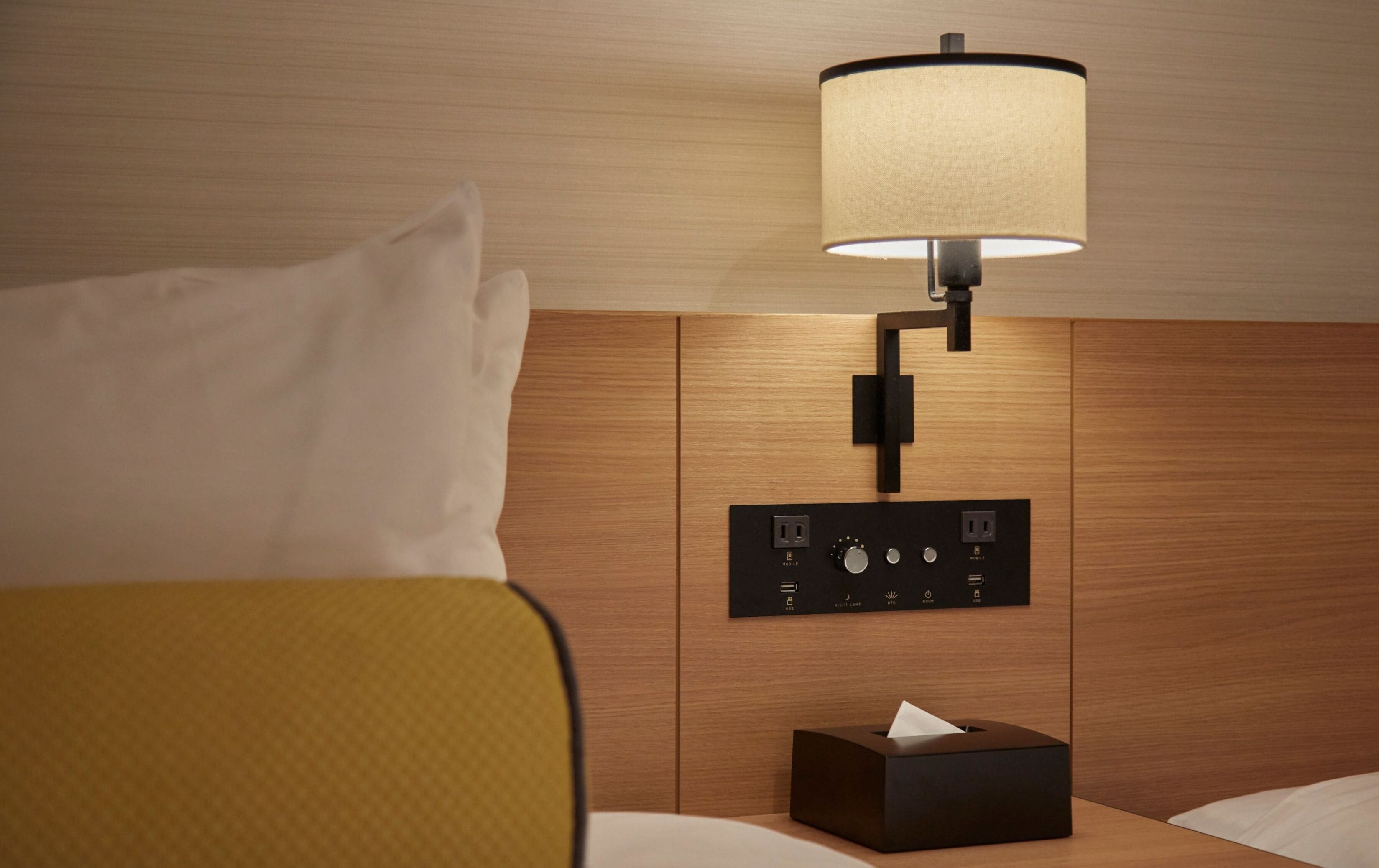 A hotel room with a bed, a bedside table with a lamp, a power outlet, and a tissue box.