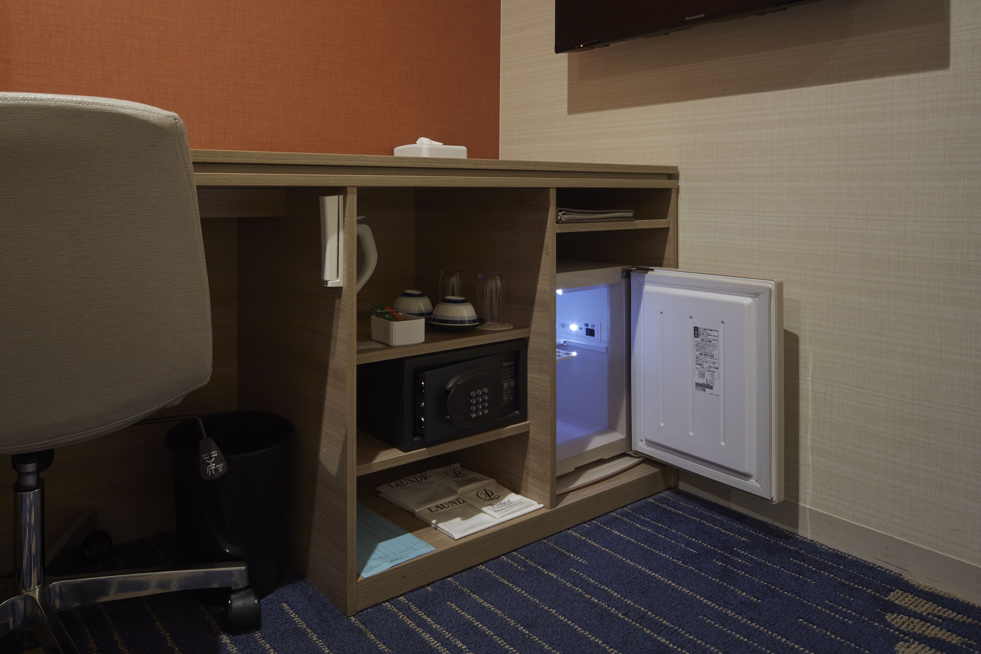 A compact hotel room with a microwave, a small refrigerator, a desk with a lamp, and a trash bin.