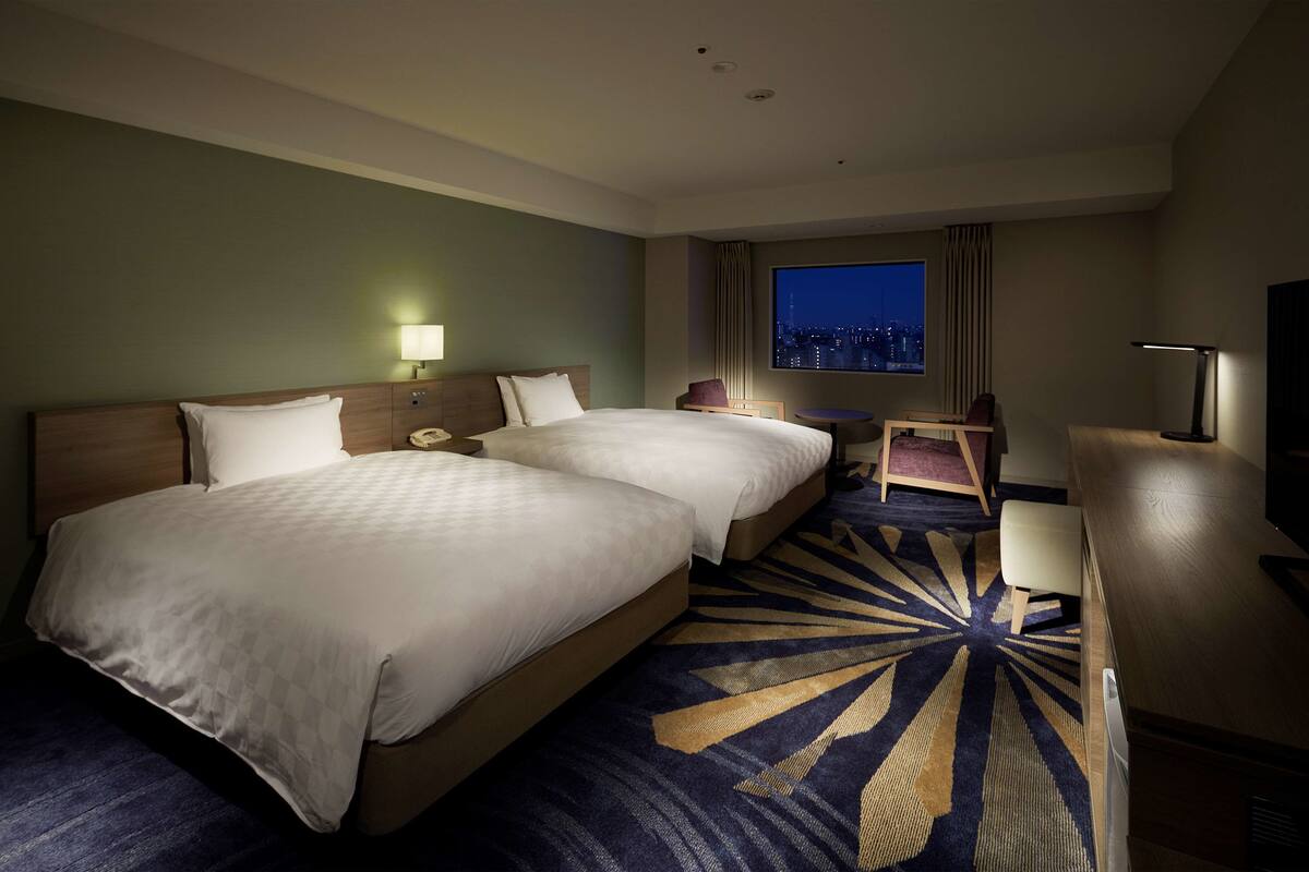 A hotel room with two beds, a desk, a chair, a TV, and a large window with a city view.