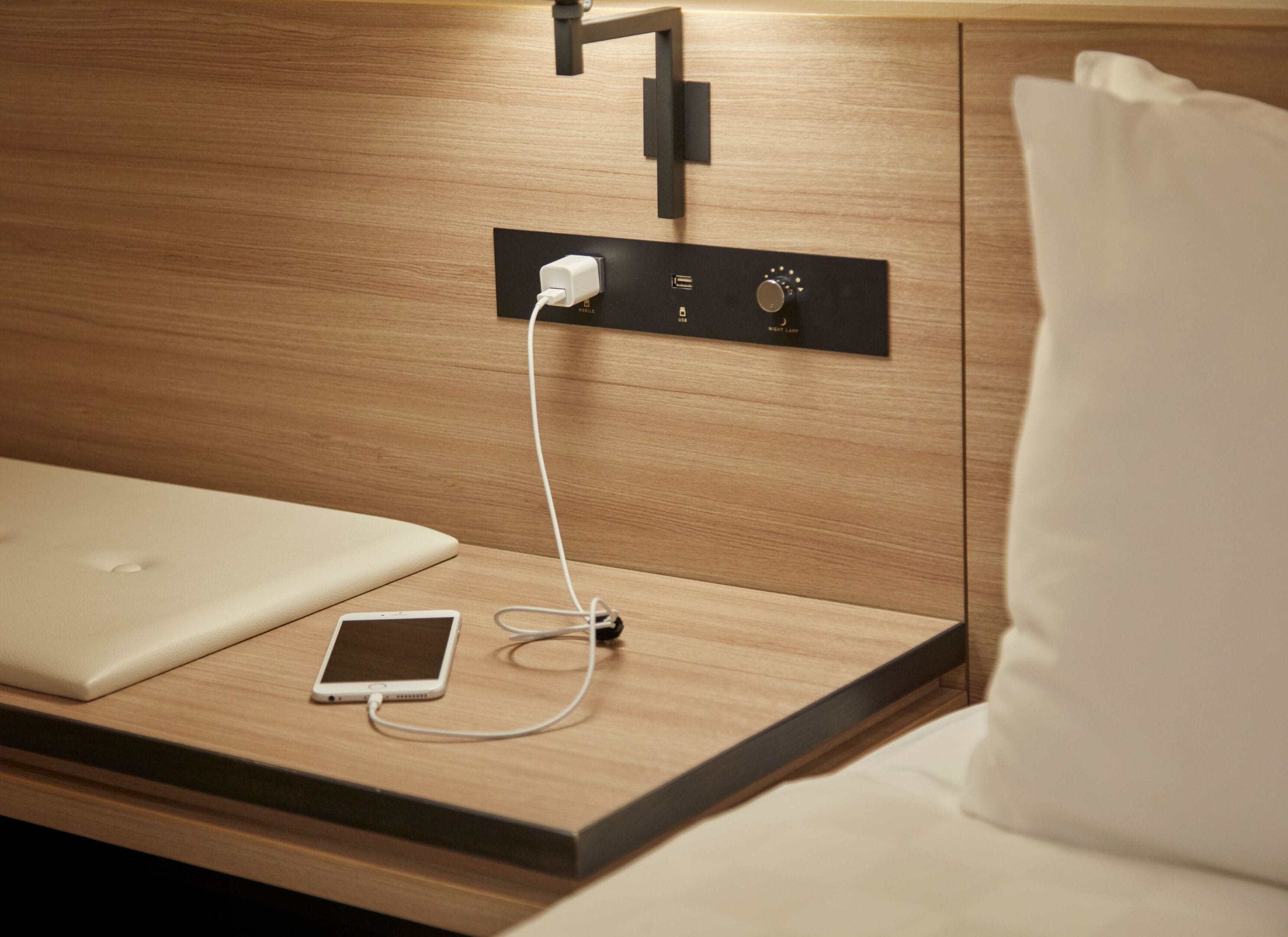 A neatly arranged hotel room with a wooden headboard, a bedside table with a laptop, a smartphone, and a power outlet.