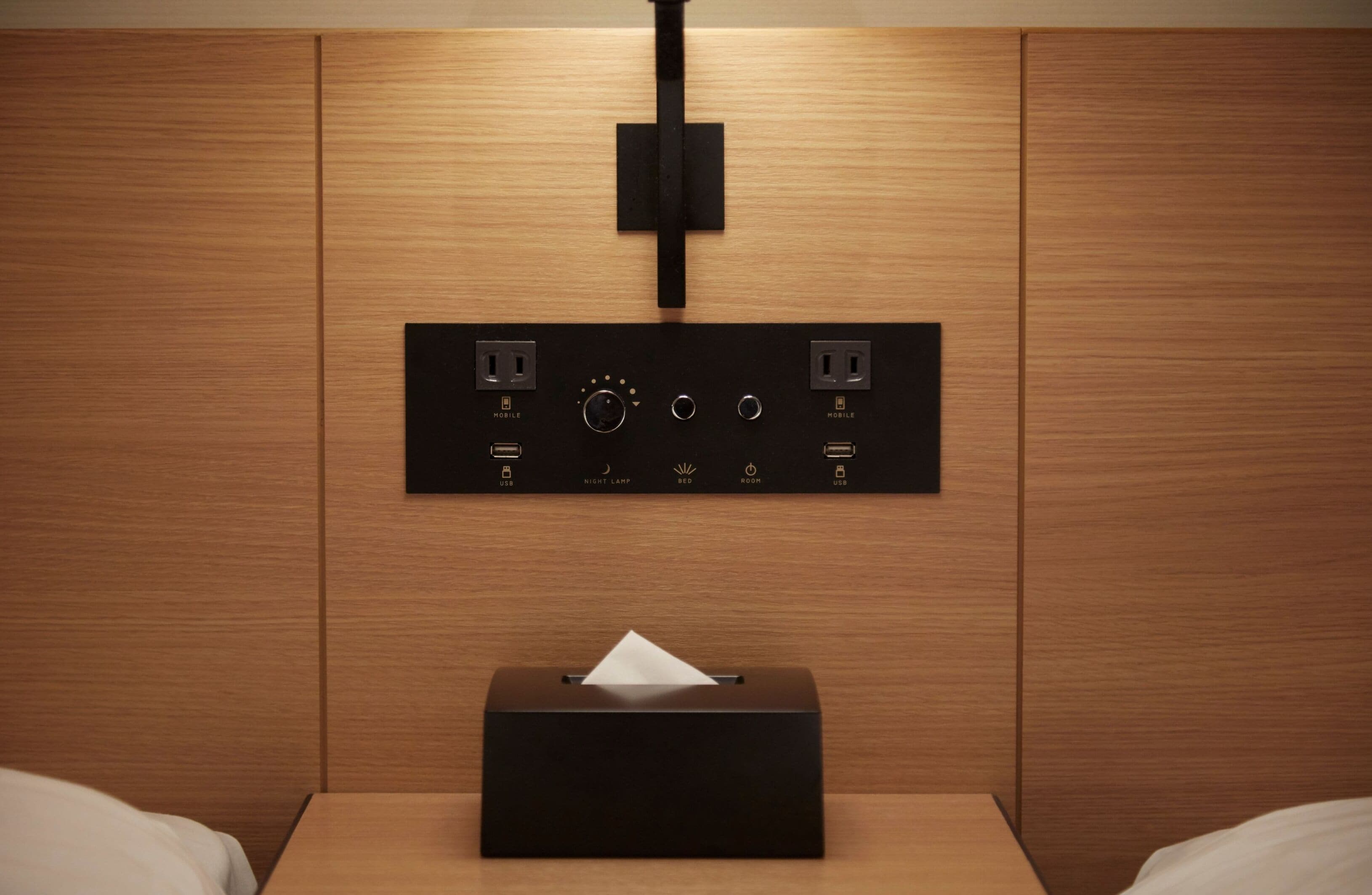 A hotel room with a wooden panel wall, a bedside table with a tissue box, and a control panel with power outlets and a light switch.
