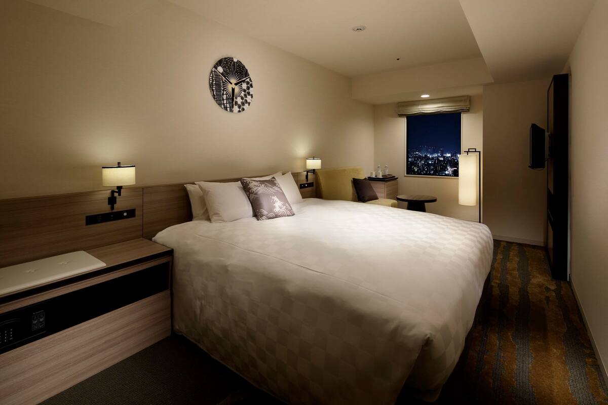 A hotel room with a large bed, bedside lamps, a wall clock, a window with a city view, and a flat-screen TV.