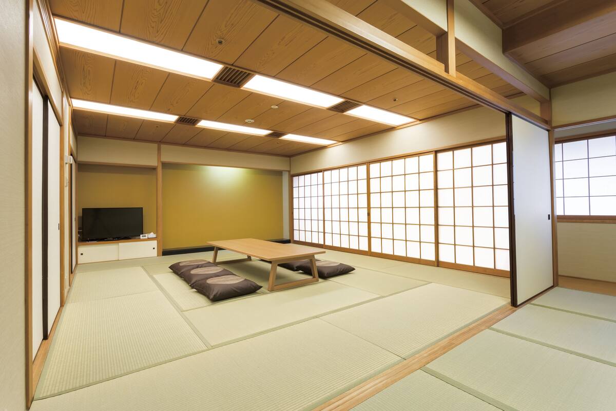 A traditional Japanese room with tatami flooring, a low table, cushions, sliding doors, and a television.
