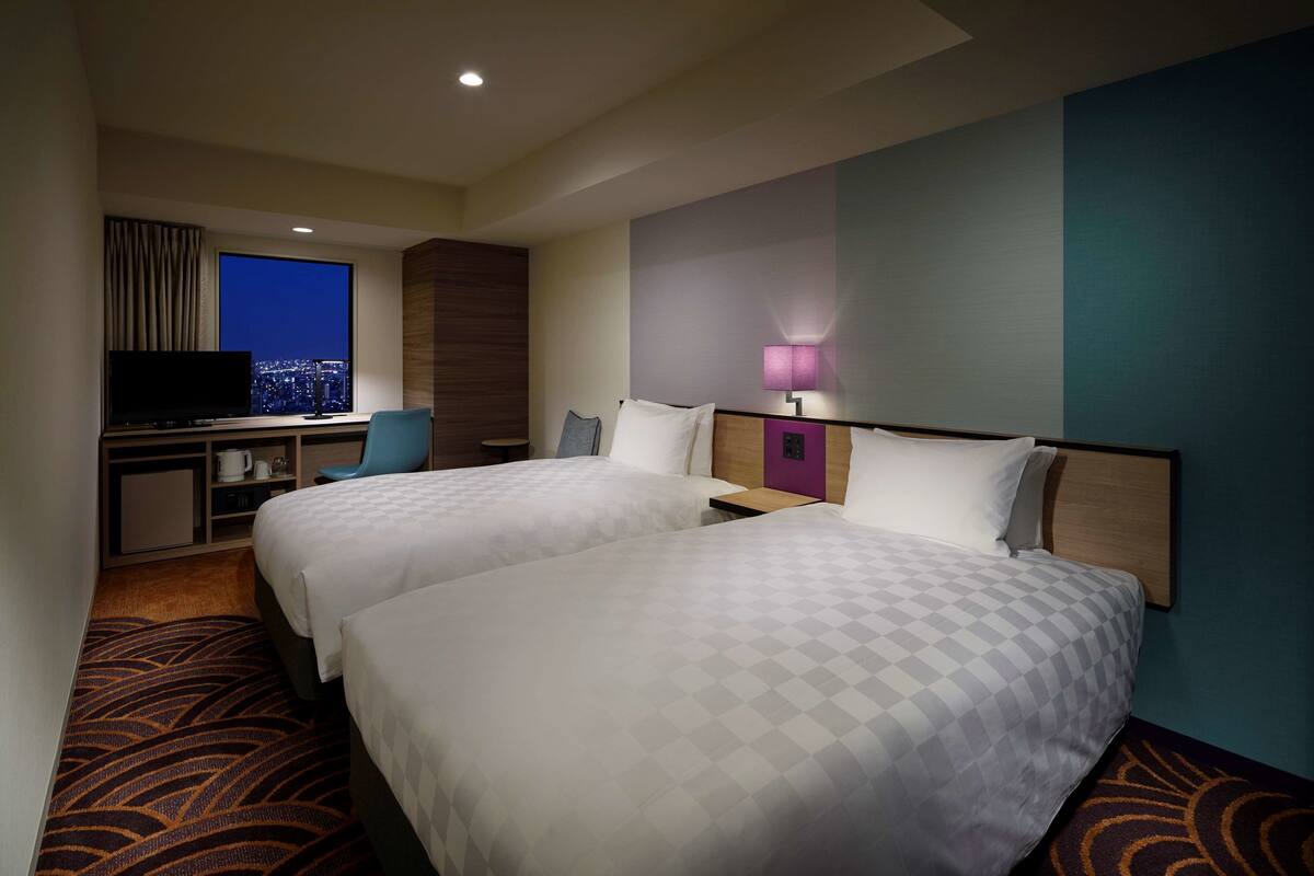 A hotel room with two beds, a TV, a desk, and a city view.