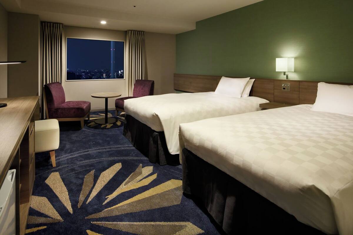 A hotel room with two beds, a desk, a chair, a small table, and a view of the city at night.