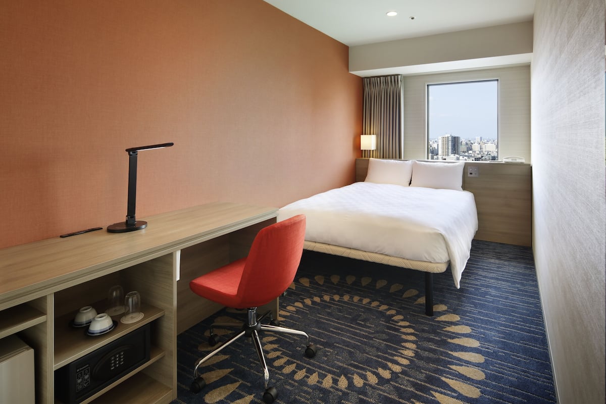 A hotel room with a bed, a desk with a chair, a microwave, and a view of the city through the window.