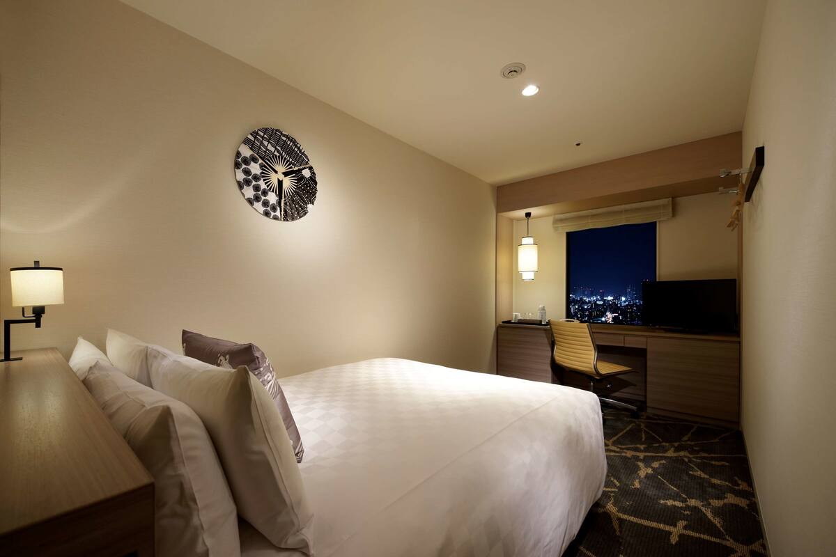 A hotel room with a large bed, a desk with a lamp, a chair, a desk lamp, a wall clock, and a view of the city at night.