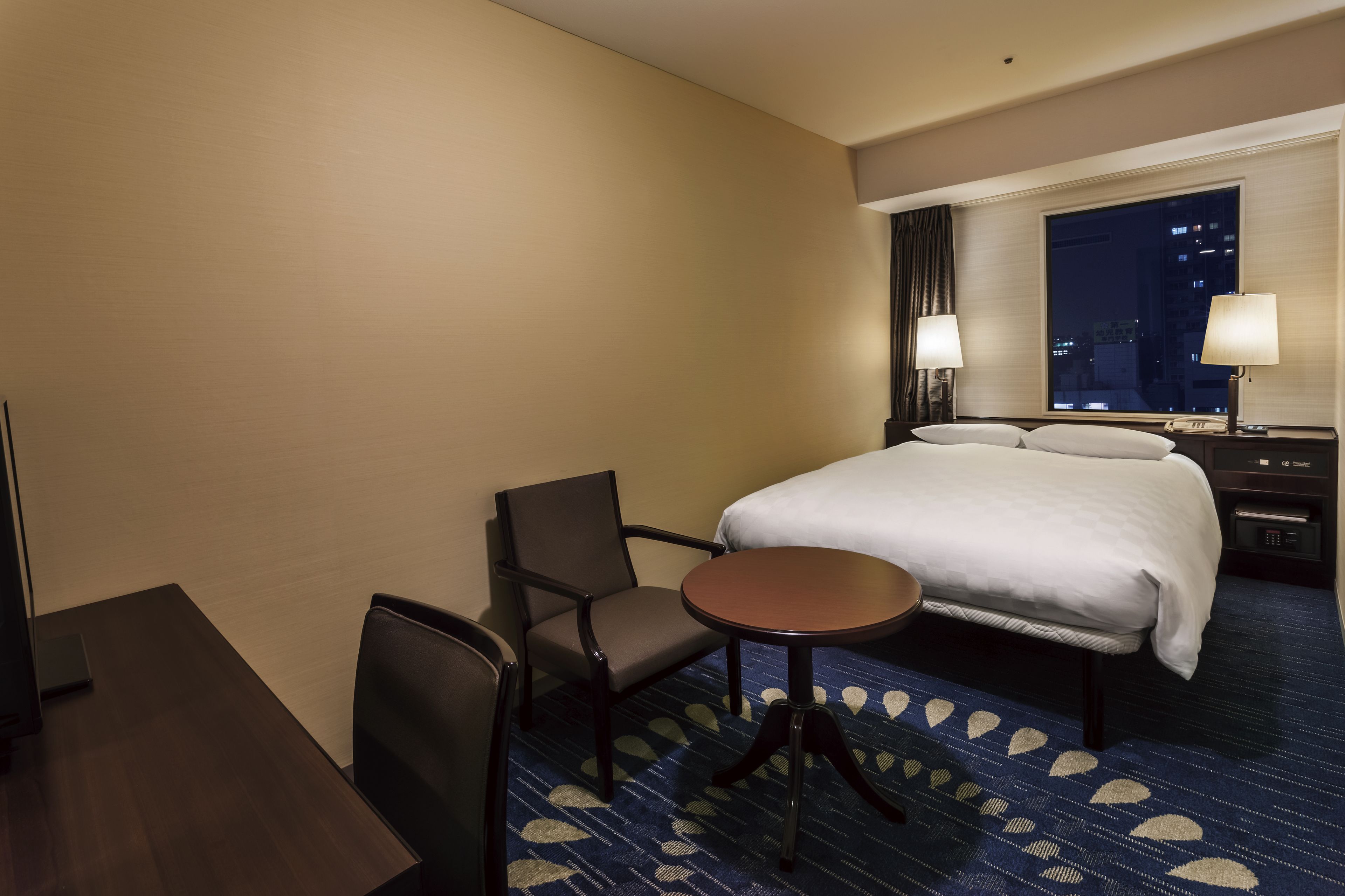 A hotel room with a bed, two chairs, a small table, a desk, and a window with a city view.