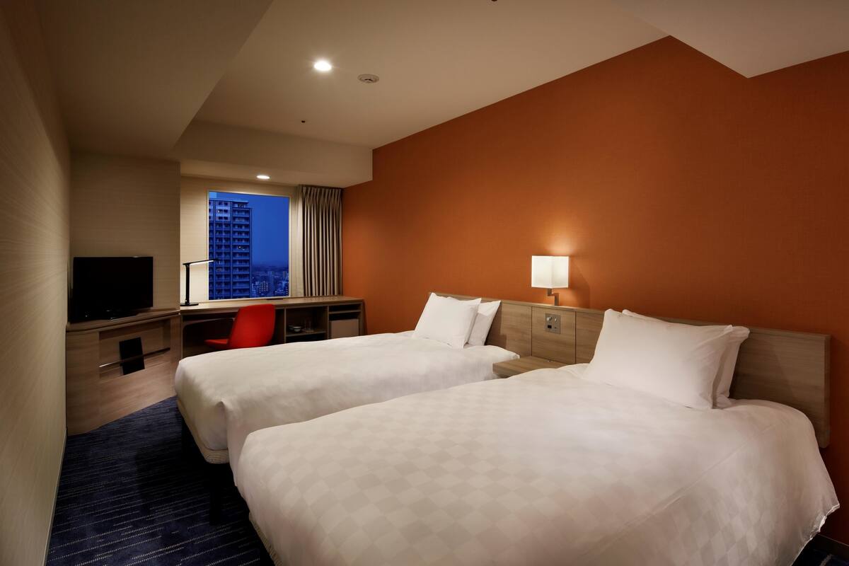 A hotel room with two beds, a desk, a TV, and a window with a city view.