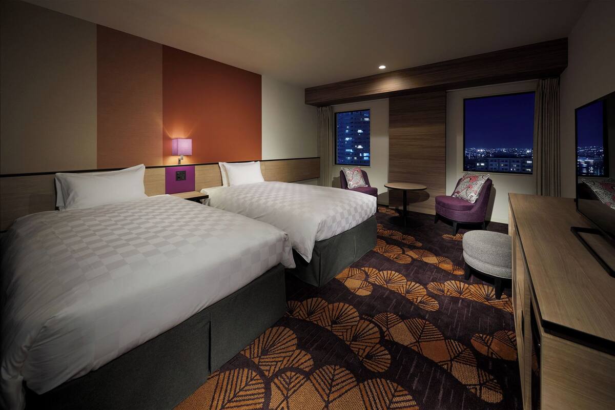 A hotel room with two beds, a small table, a chair, and a view of the city at night.