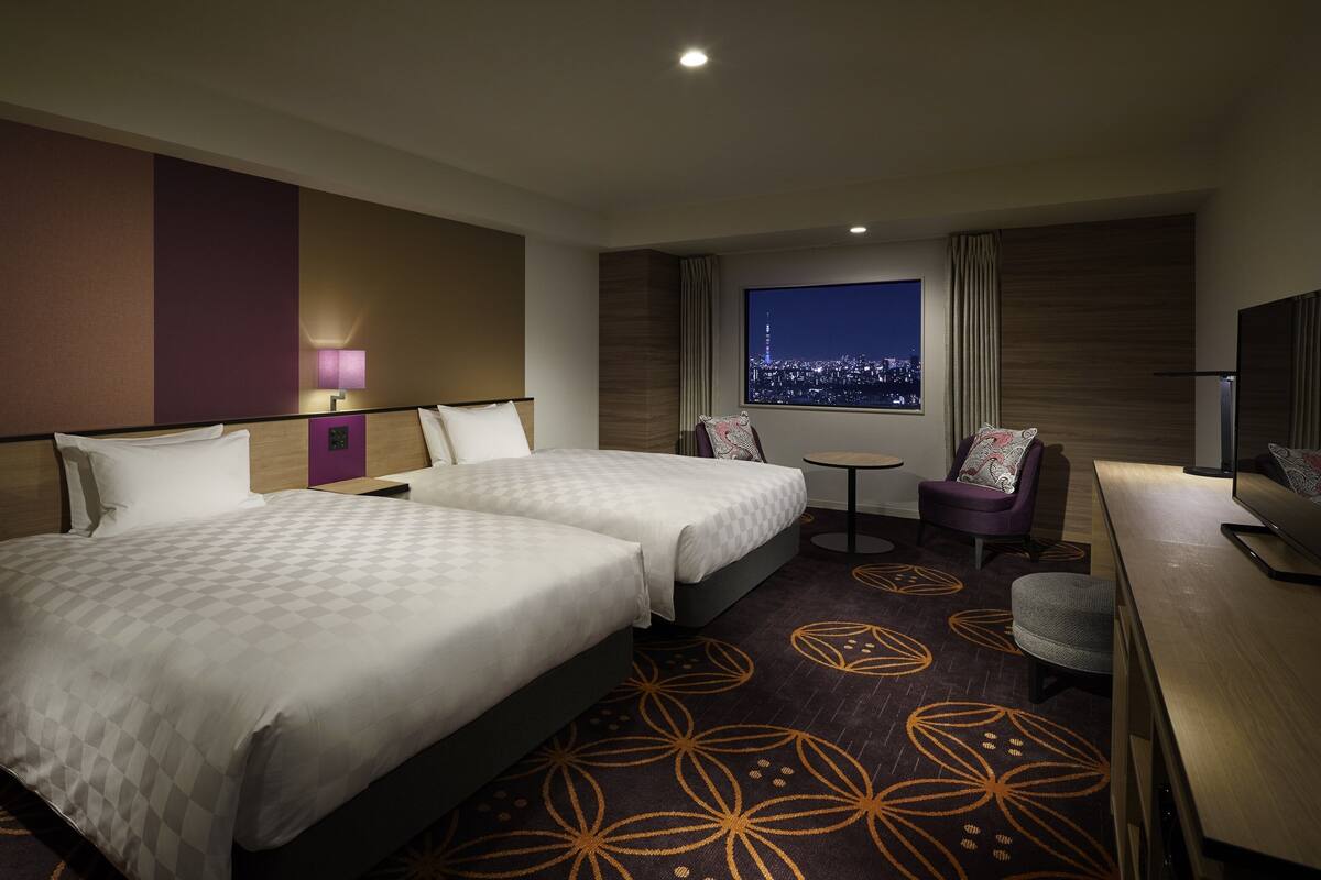 A hotel room with two beds, a small table, a chair, a TV, and a city view.