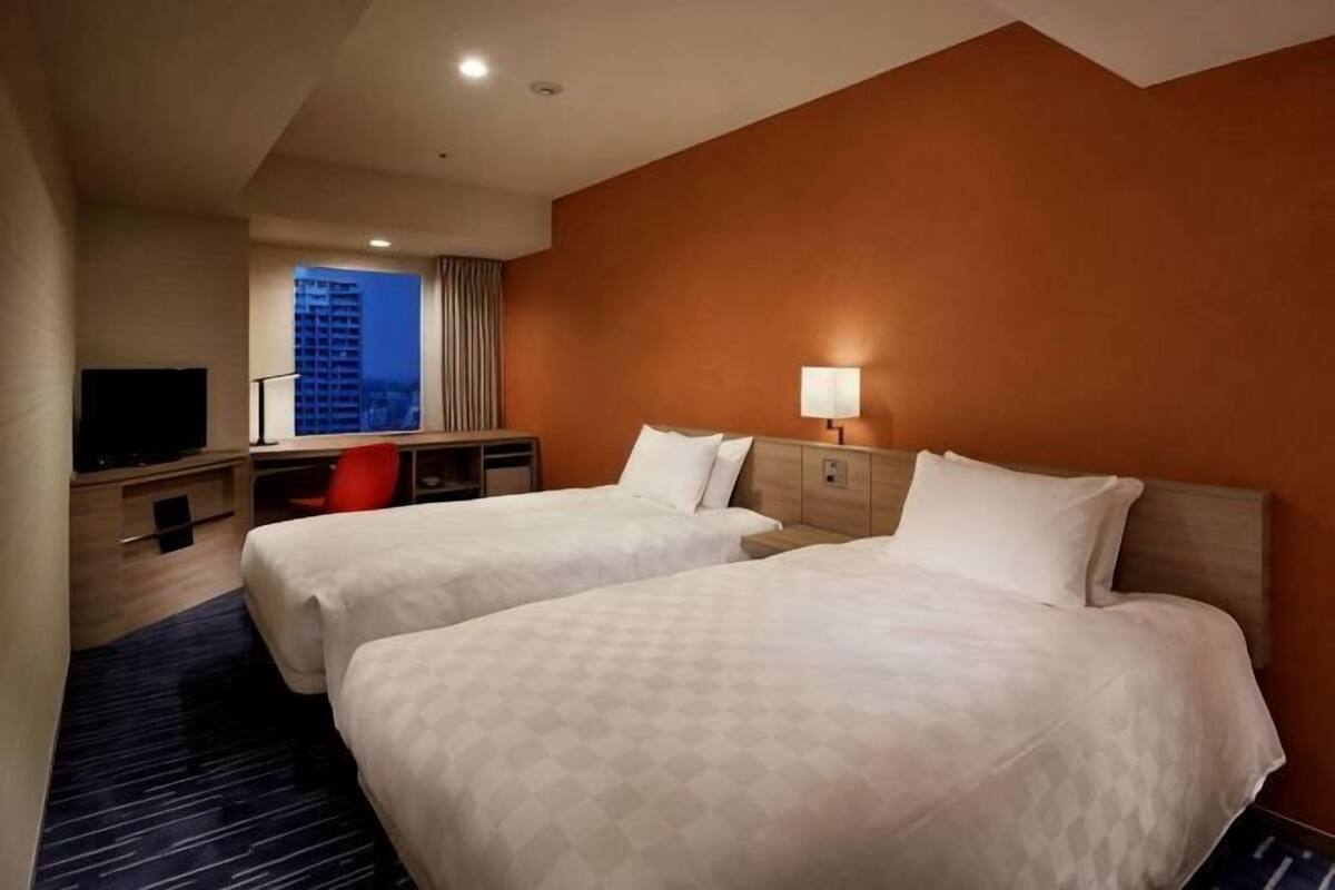 A hotel room with two beds, a desk with a chair, a television, and a window with a view of a building.