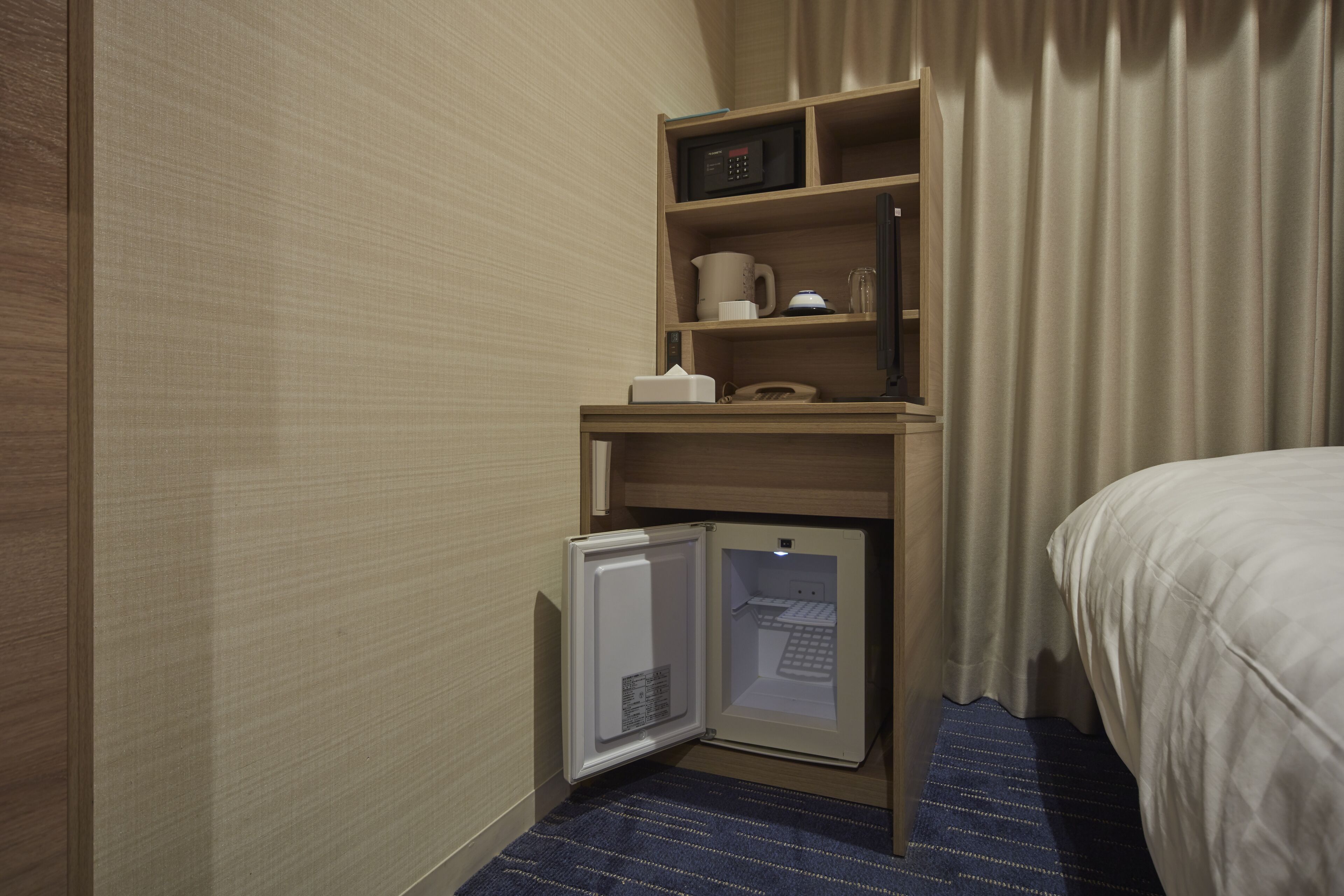 A compact hotel room with a built-in minibar, a small refrigerator, a microwave, and a bedside table with a lamp.