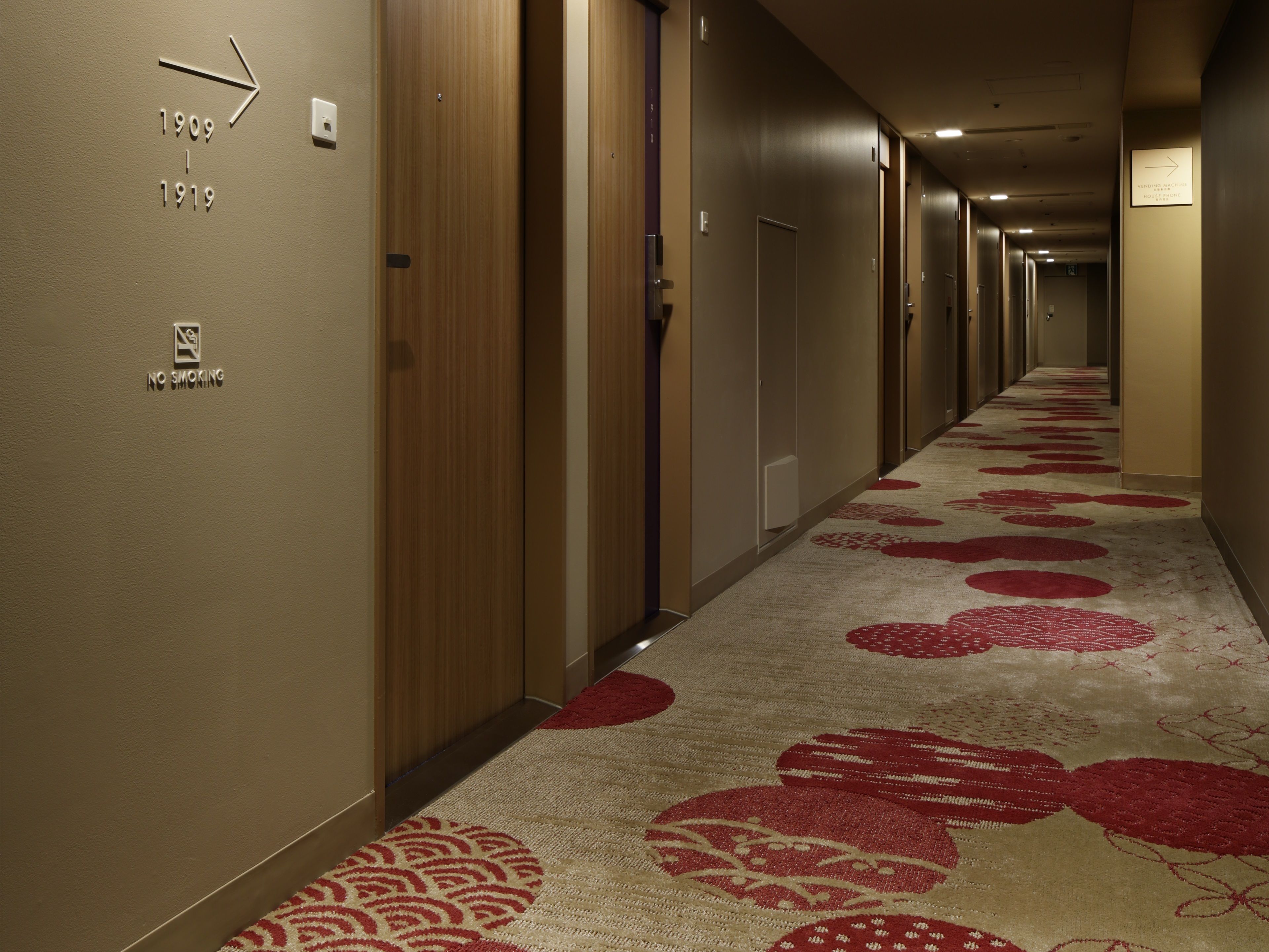 A hotel hallway with a carpet featuring red circular patterns, doors on the left, and a sign indicating the vending machine location.