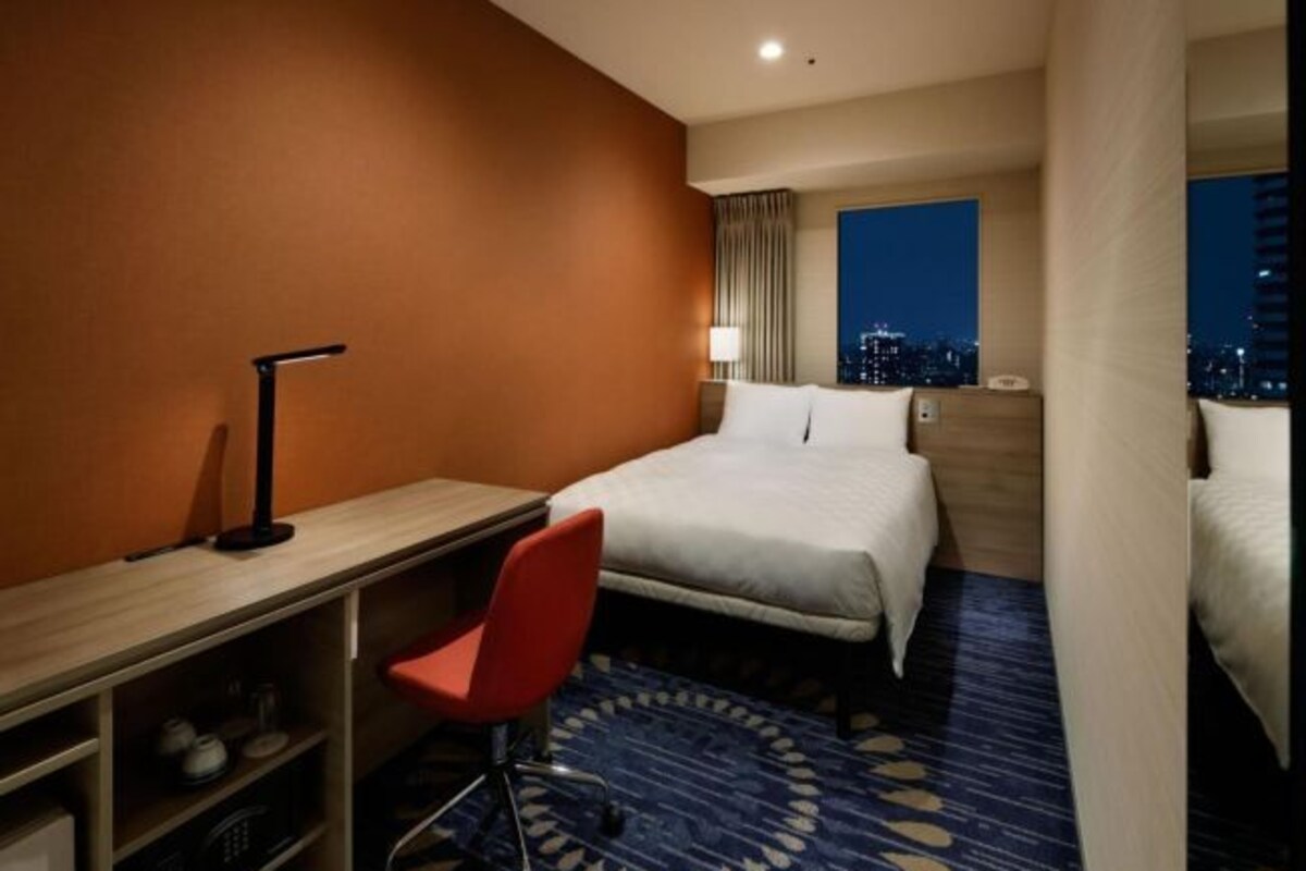 A hotel room with a bed, a desk with a chair, a lamp, and a view of the city at night.