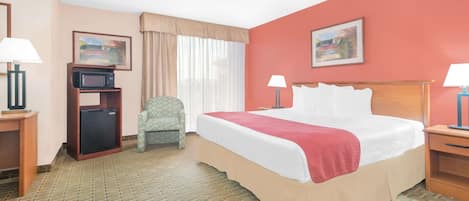 A hotel room with a large bed, a desk with a computer, a chair, a microwave, and a nightstand with a lamp.