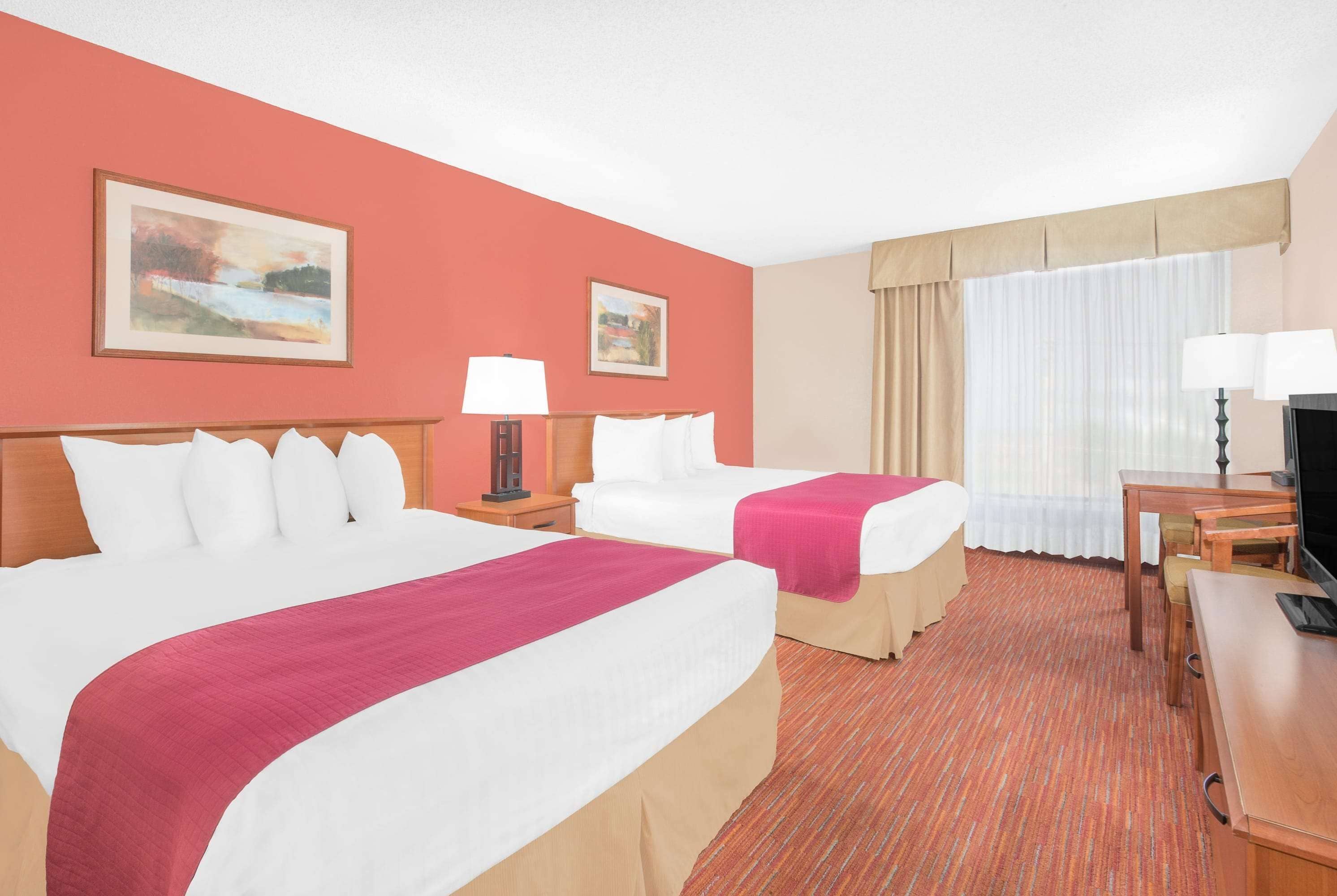 A hotel room with two beds, a desk, a chair, a television, and two paintings on the wall.