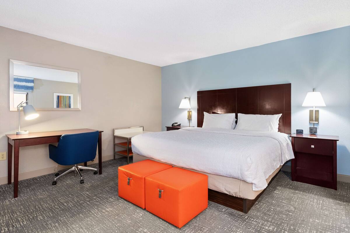 A hotel room with a large bed, a desk with a chair, two orange ottomans, a nightstand with a lamp, and a wall-mounted mirror.