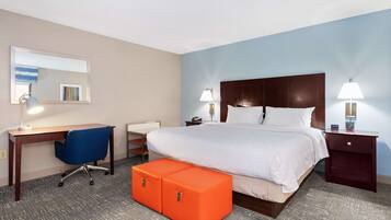Quarto de hotel com uma cama grande, uma escrivaninha com cadeira, dois pufes laranja, uma mesinha de cabeceira com abajur e um espelho fixado na parede.