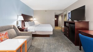 A hotel room with a bed, a sofa, a desk, a TV, and a minibar.