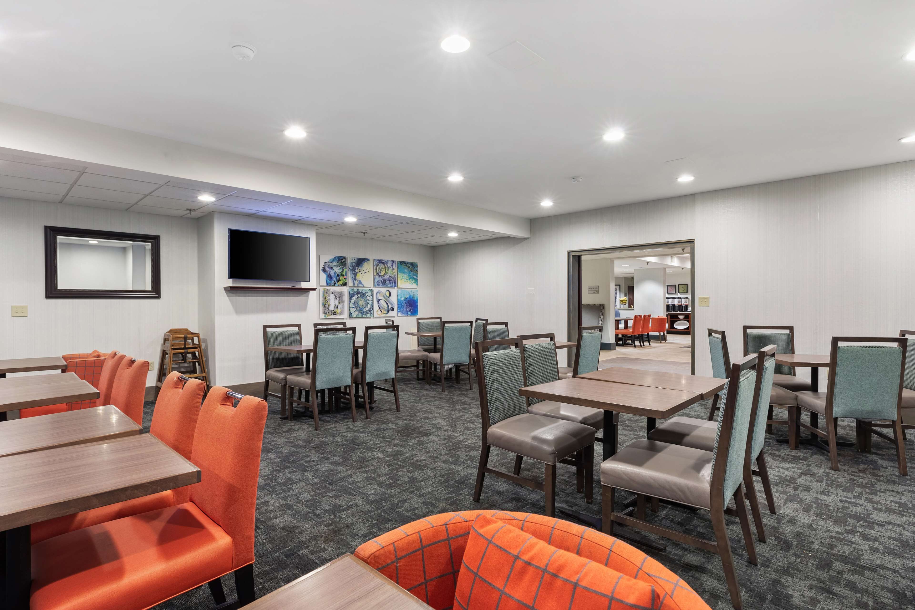 A hotel dining area with multiple tables and chairs, a flat-screen TV mounted on the wall, and artwork on the back wall.
