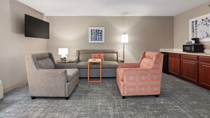 A living room with a sofa, two armchairs, a coffee table, a flat-screen TV, a lamp, and a kitchen area with cabinets and a microwave. - Hampton Inn Rock Hill (Rock Hill)