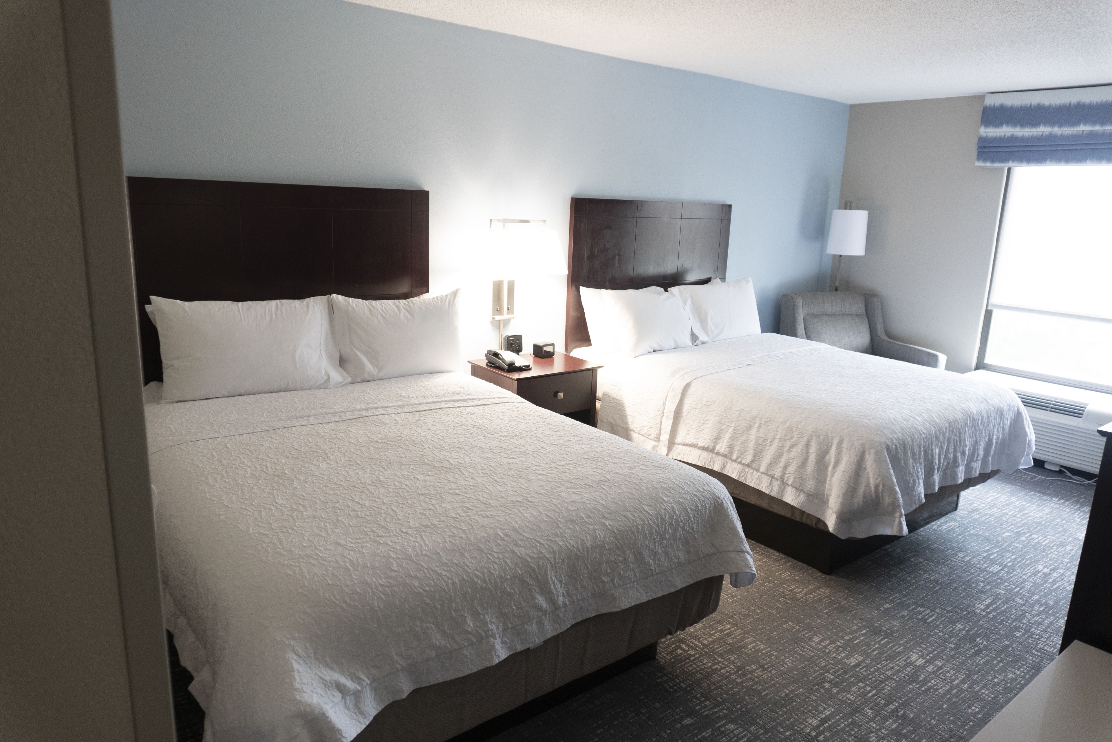 Een hotelkamer met twee bedden, een nachtkastje met een lamp, een stoel en een groot raam.