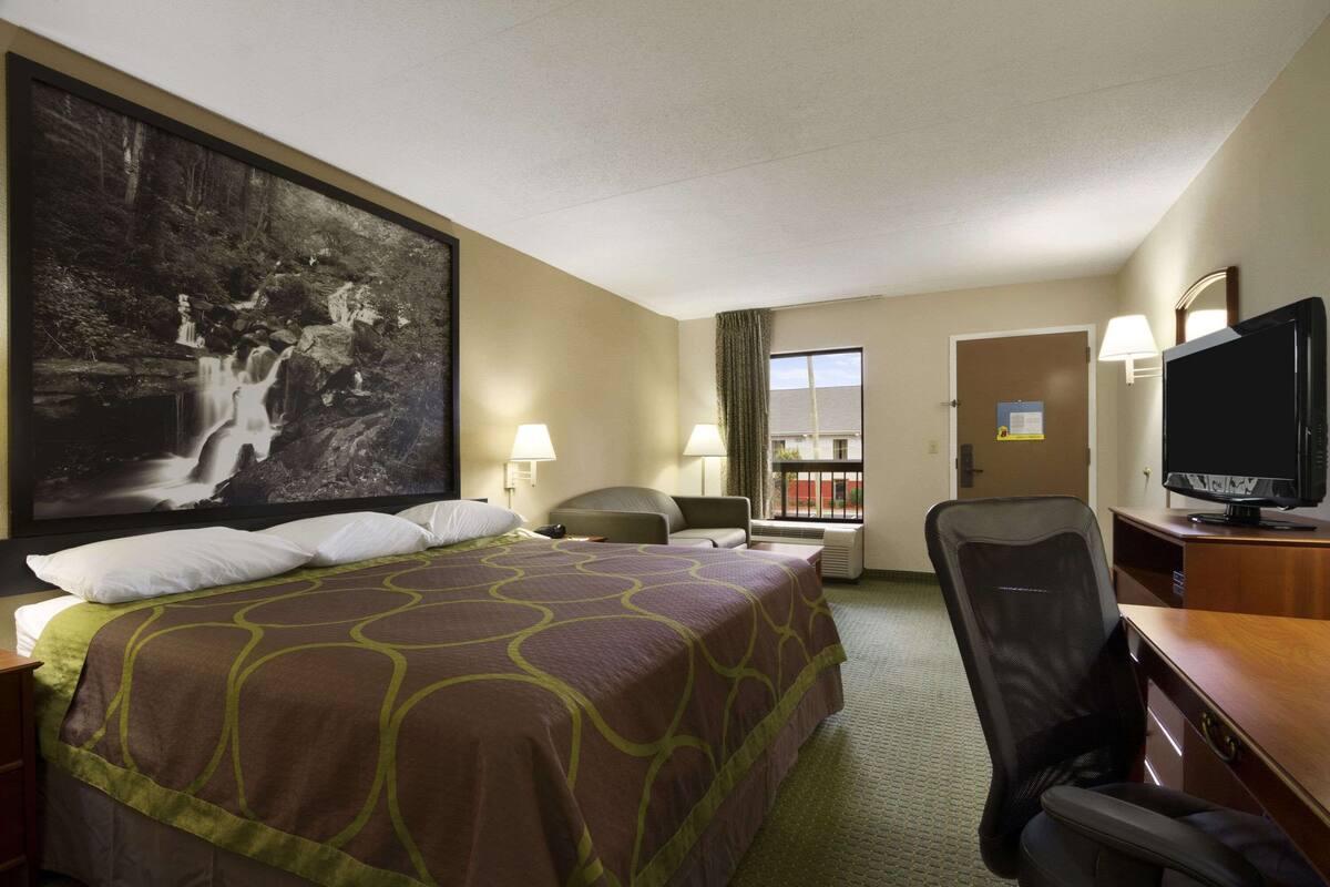 Quarto de hotel com uma cama grande, uma escrivaninha com televisão, uma cadeira e janela com vista para um edifício.