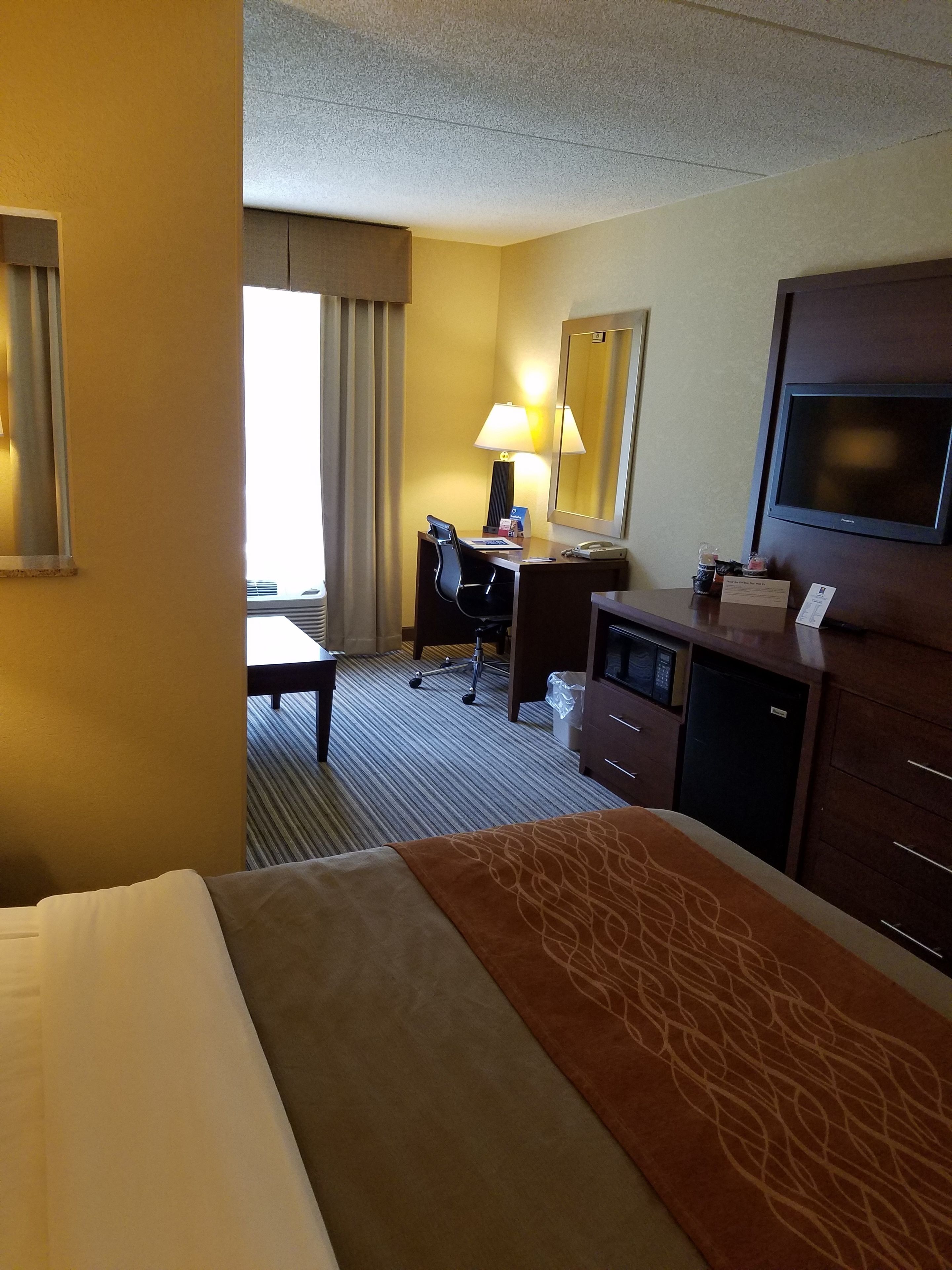 A hotel room with a bed, a desk with a chair, a lamp, a television, and a minibar.