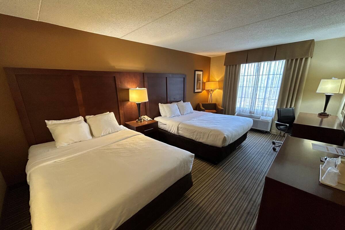 A hotel room with two beds, a desk, a chair, a lamp, and a window with curtains.