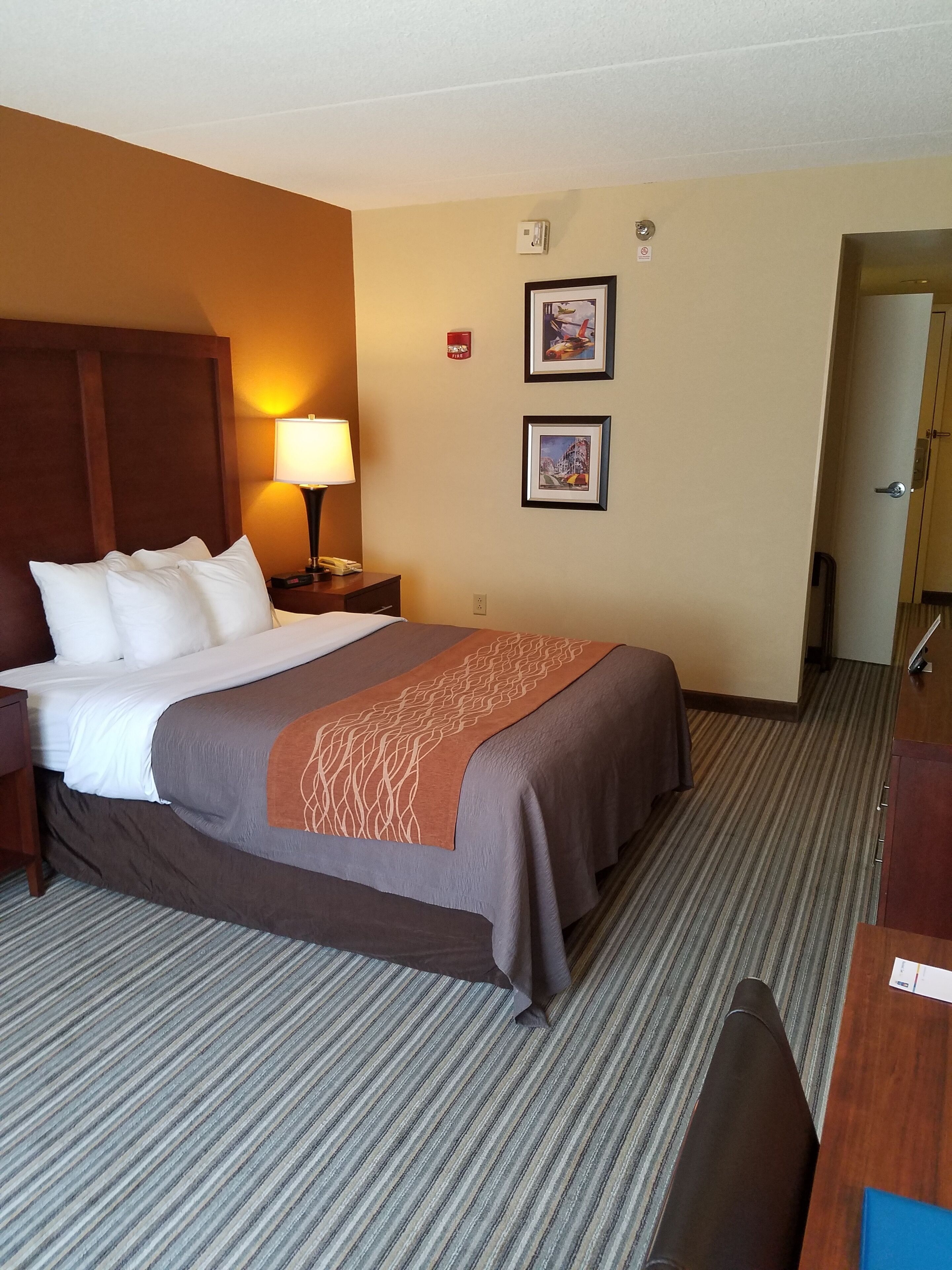 A hotel room with a bed, a nightstand, a lamp, two framed pictures on the wall, and a door leading to another room.