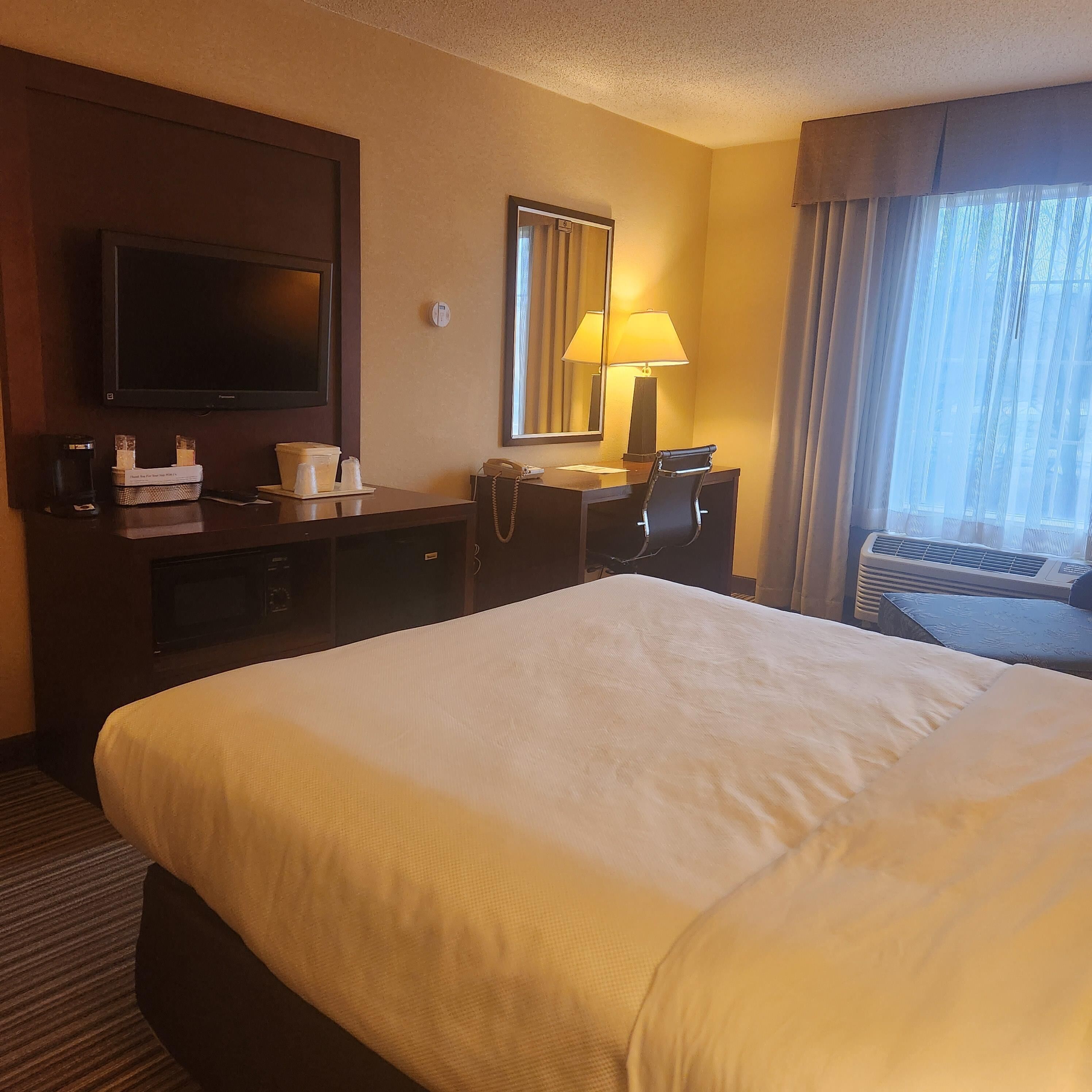 A hotel room with a large bed, a desk with a lamp, a television, and a window with curtains.