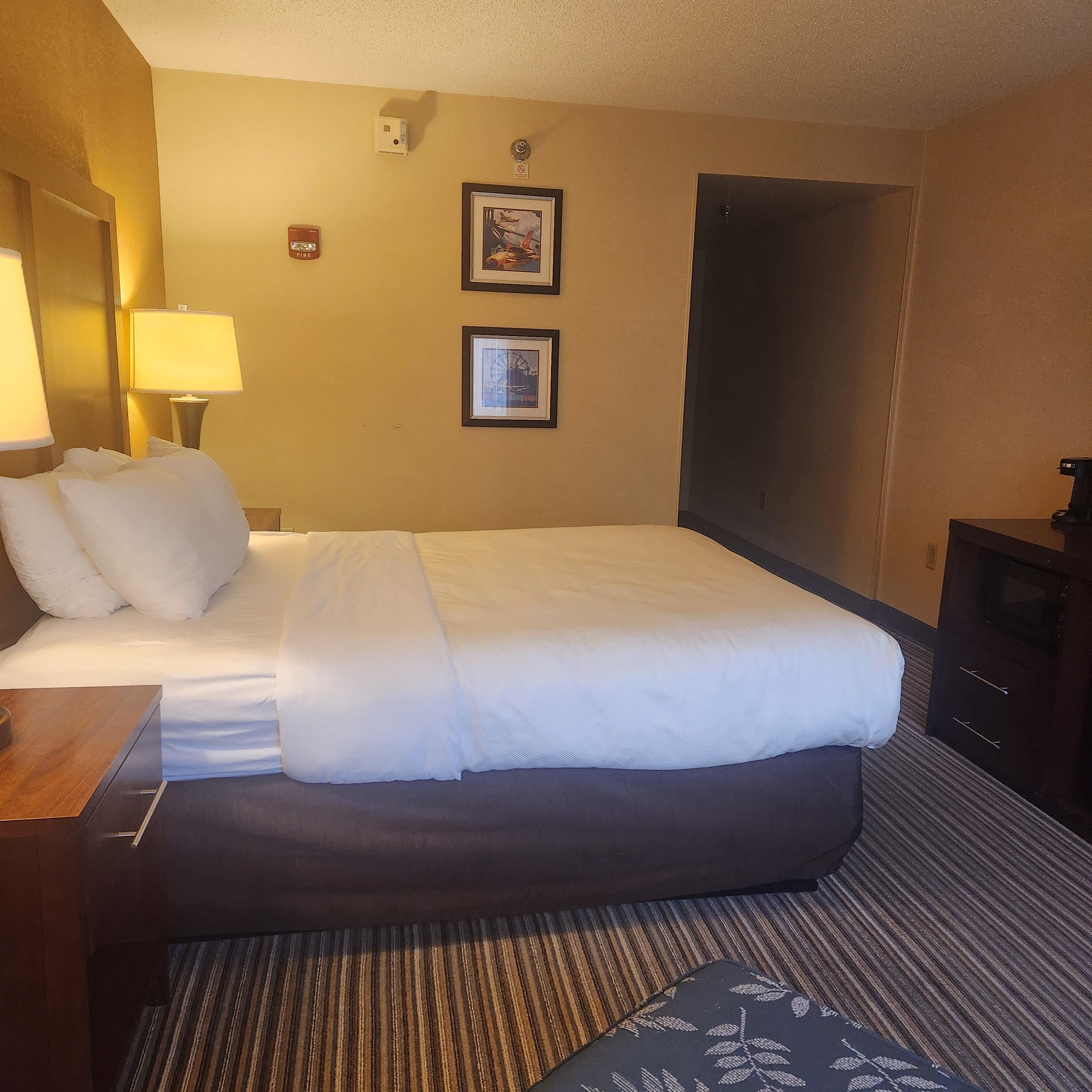 A hotel room with a large bed, a bedside table with a lamp, two framed pictures on the wall, and a dark-colored wardrobe.