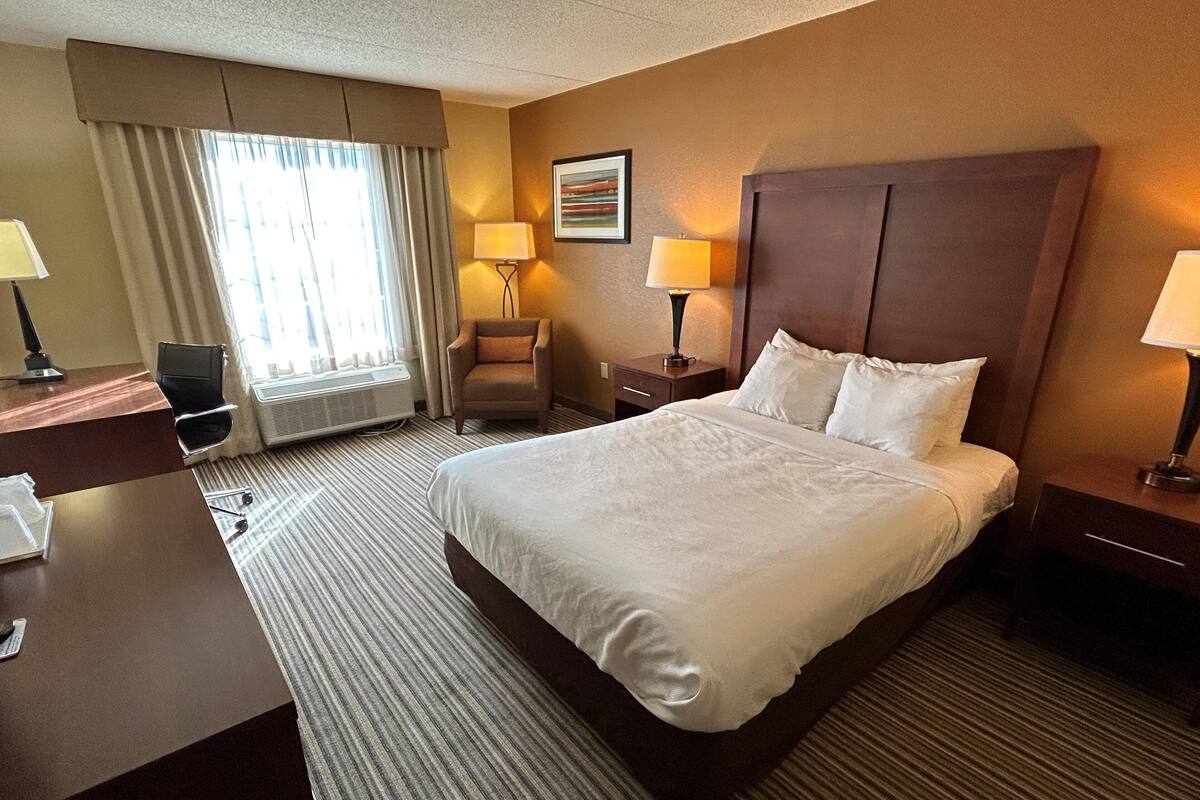 A hotel room with a large bed, a desk with a chair, two bedside lamps, a chair, and a window with curtains.