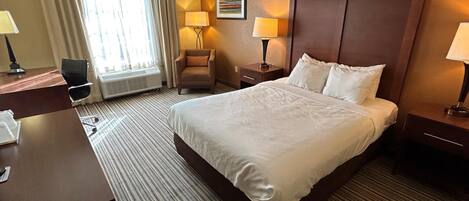 A hotel room with a large bed, a desk with a chair, two bedside lamps, a chair, and a window with curtains.