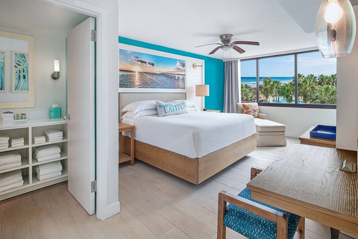 A modern bedroom with a large bed, a ceiling fan, a view of the ocean, and a wooden table with a chair.