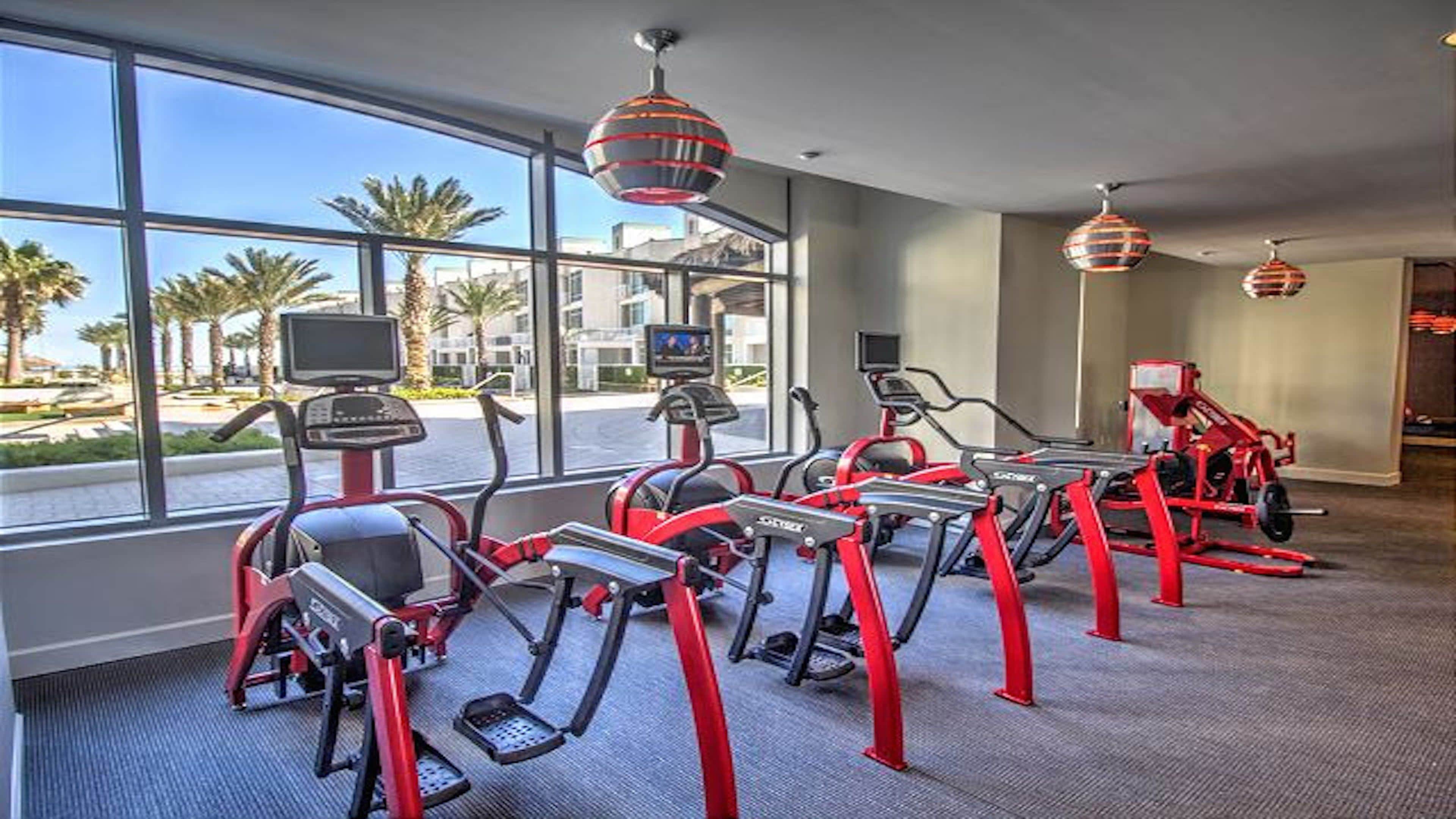 A fitness room with treadmills, exercise bikes, and a large window with a view of palm trees and buildings.