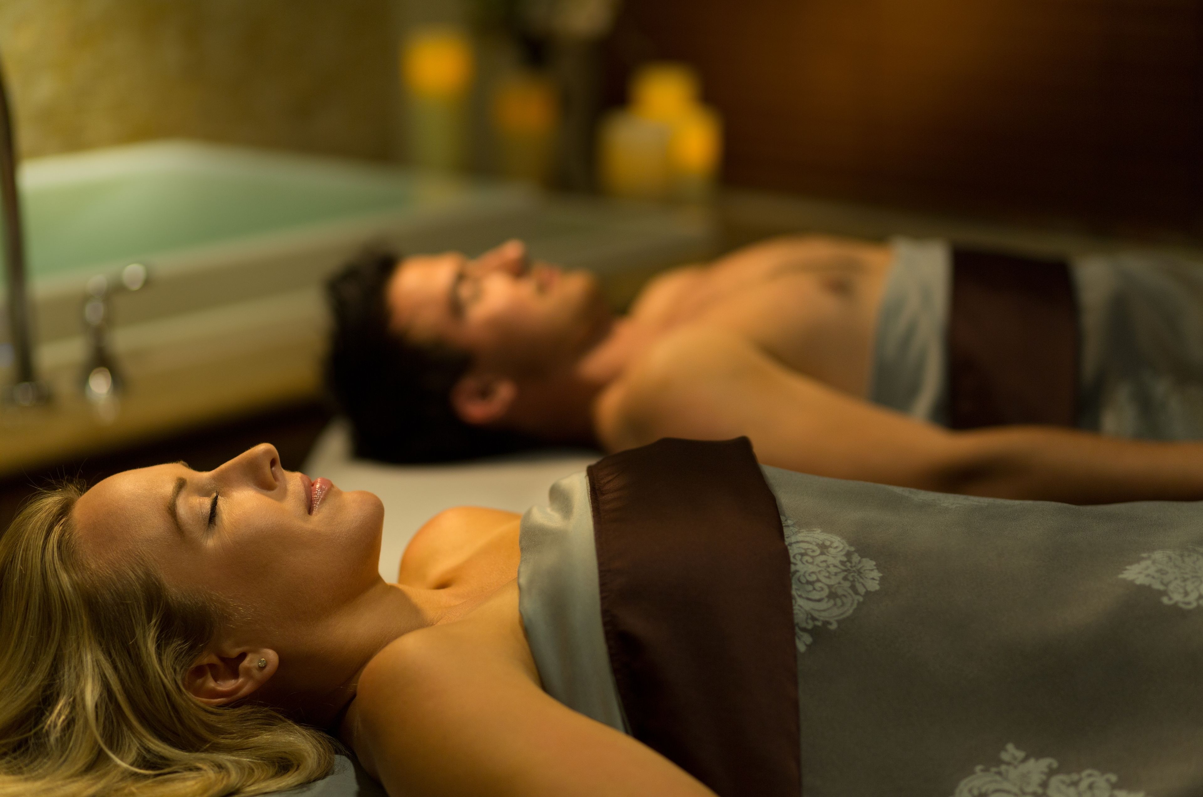Una pareja recostada en un baño de spa con una bañera visible y un sillón de masajes instalado en la pared.
