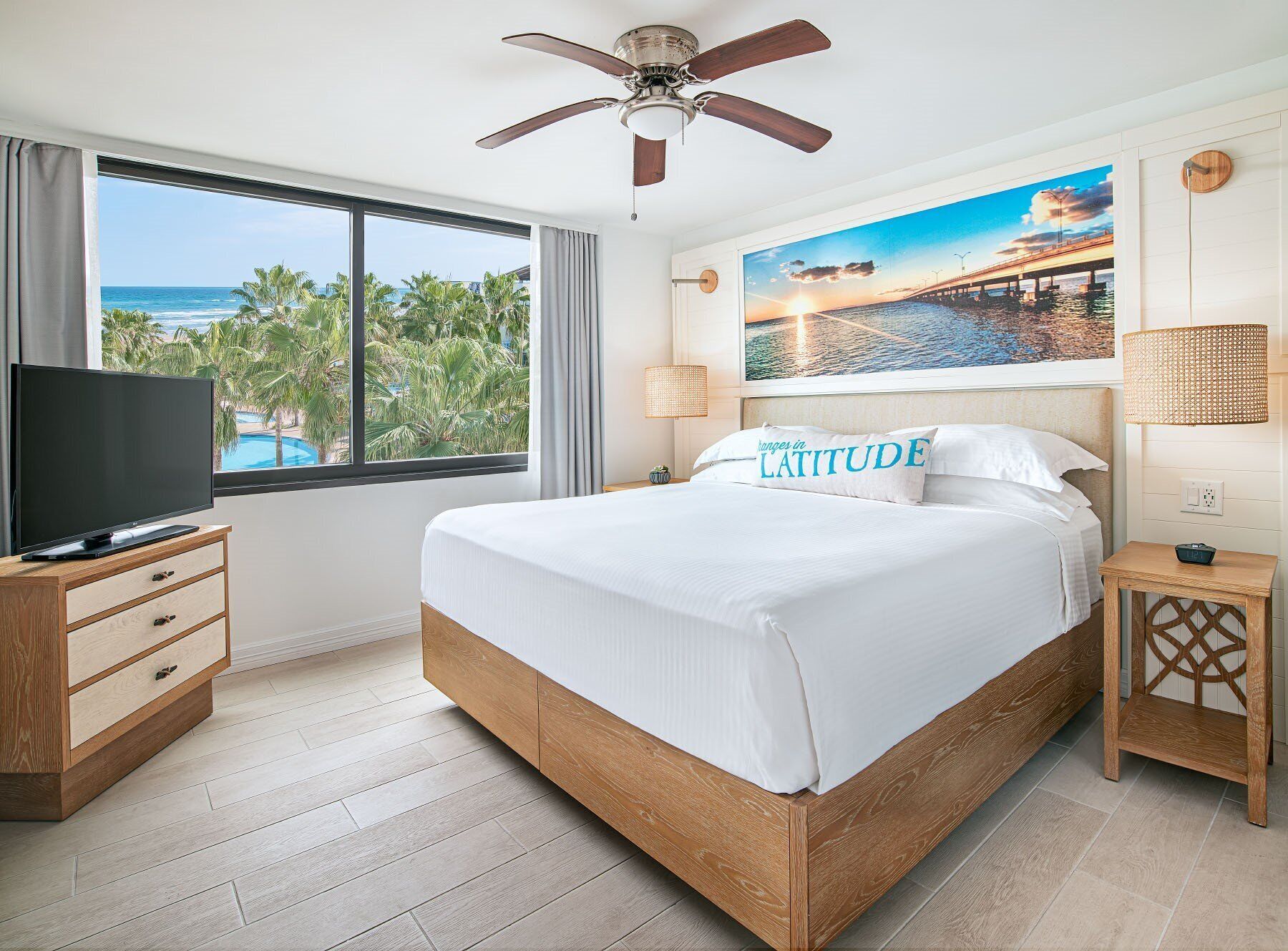 A hotel room with a large bed, a television on a wooden nightstand, a ceiling fan, a painting of a sunset over a bridge, and a view of palm trees and the ocean through a large window.
