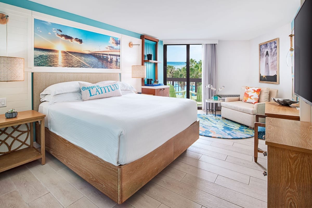 A hotel room with a large bed, a balcony with a view of the ocean, a desk with a chair, and a TV.