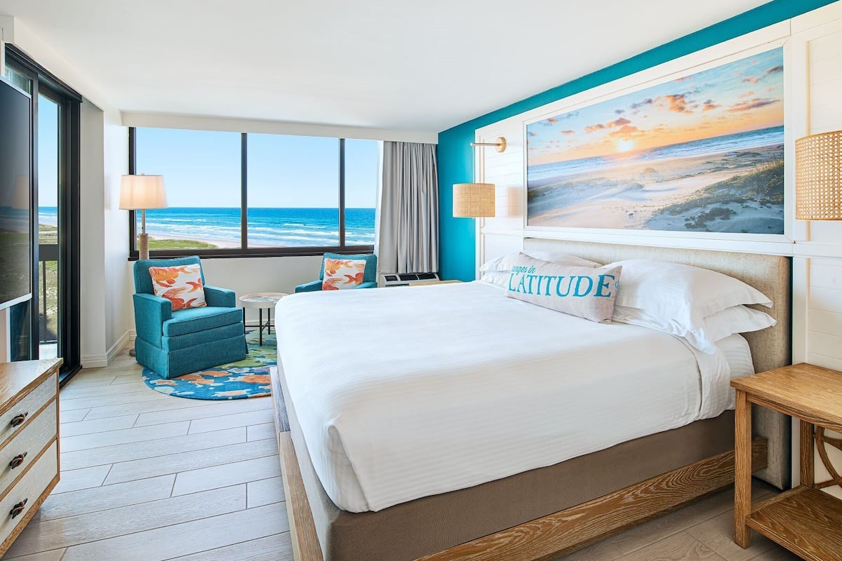 A hotel room with a large bed, a view of the ocean, a painting of a beach scene, and a blue accent wall.