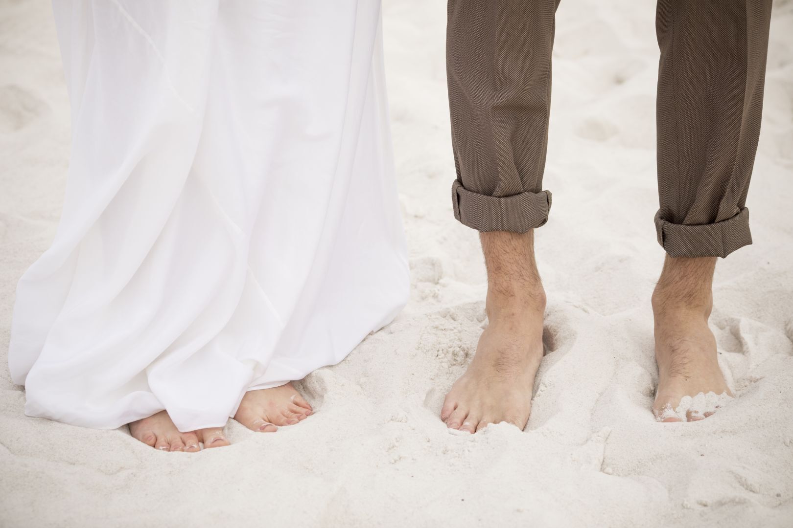 Une personne vêtue de blanc et une autre portant un pantalon marron se tiennent sur une plage de sable.