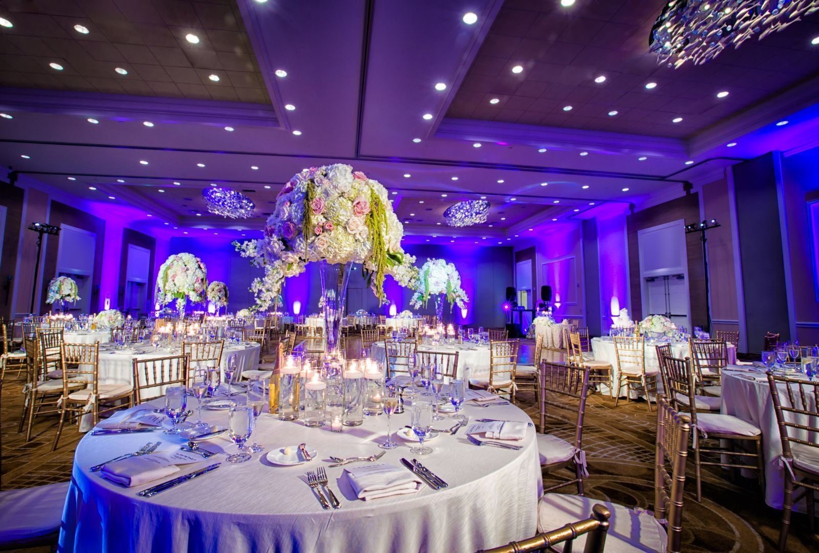 Un salón de banquetes con mesas redondas, centros de mesa florales e iluminación morada en el techo.