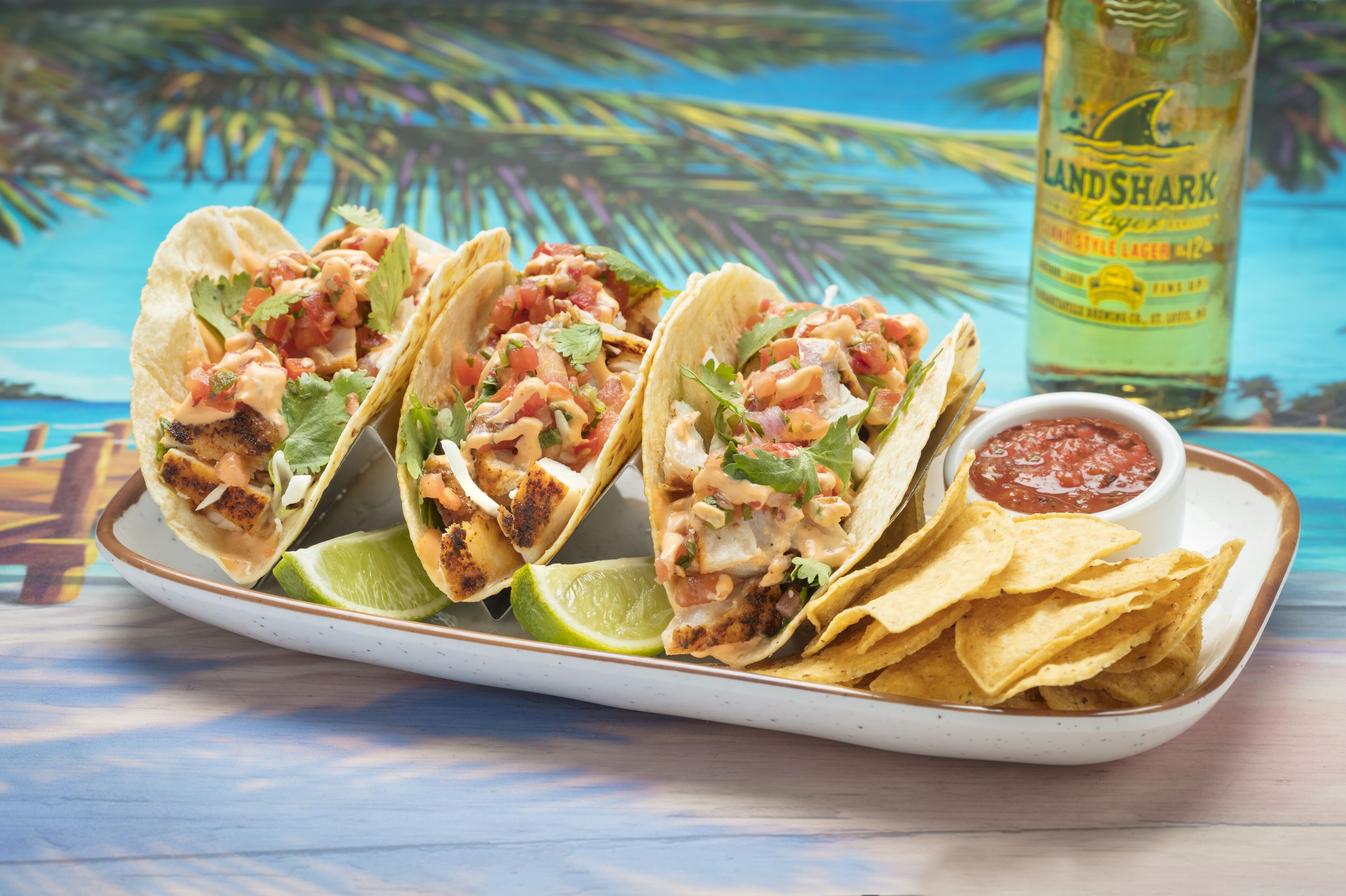 Tres tacos acompañados de chips y salsa, junto con una botella de cerveza Landshark sobre una mesa.