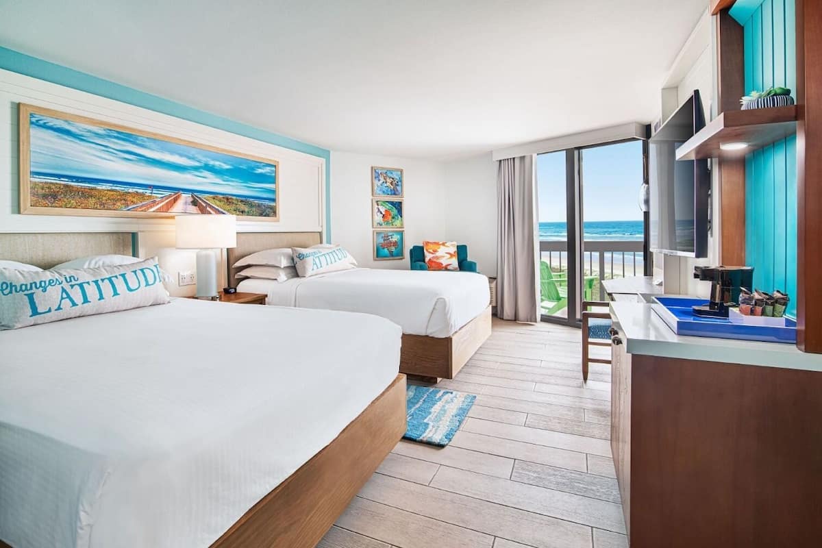 A hotel room with two beds, a balcony with a view of the ocean, and a desk with a coffee machine.