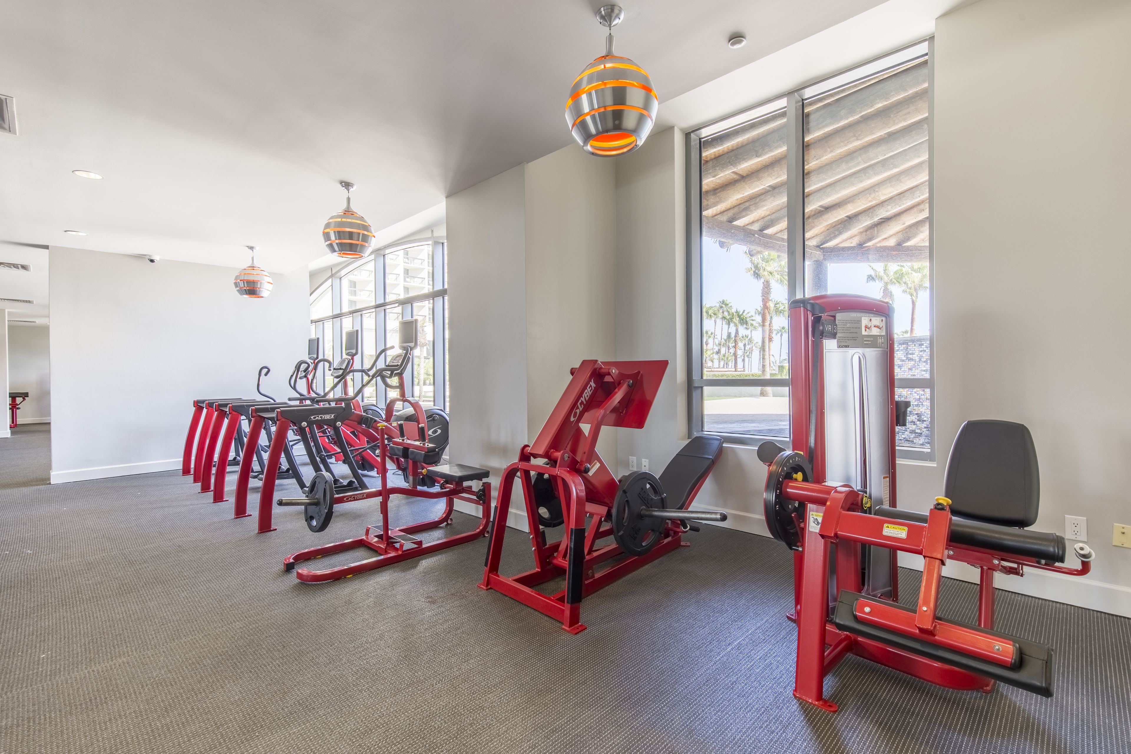 A gym with red exercise equipment, large windows, and hanging pendant lights.